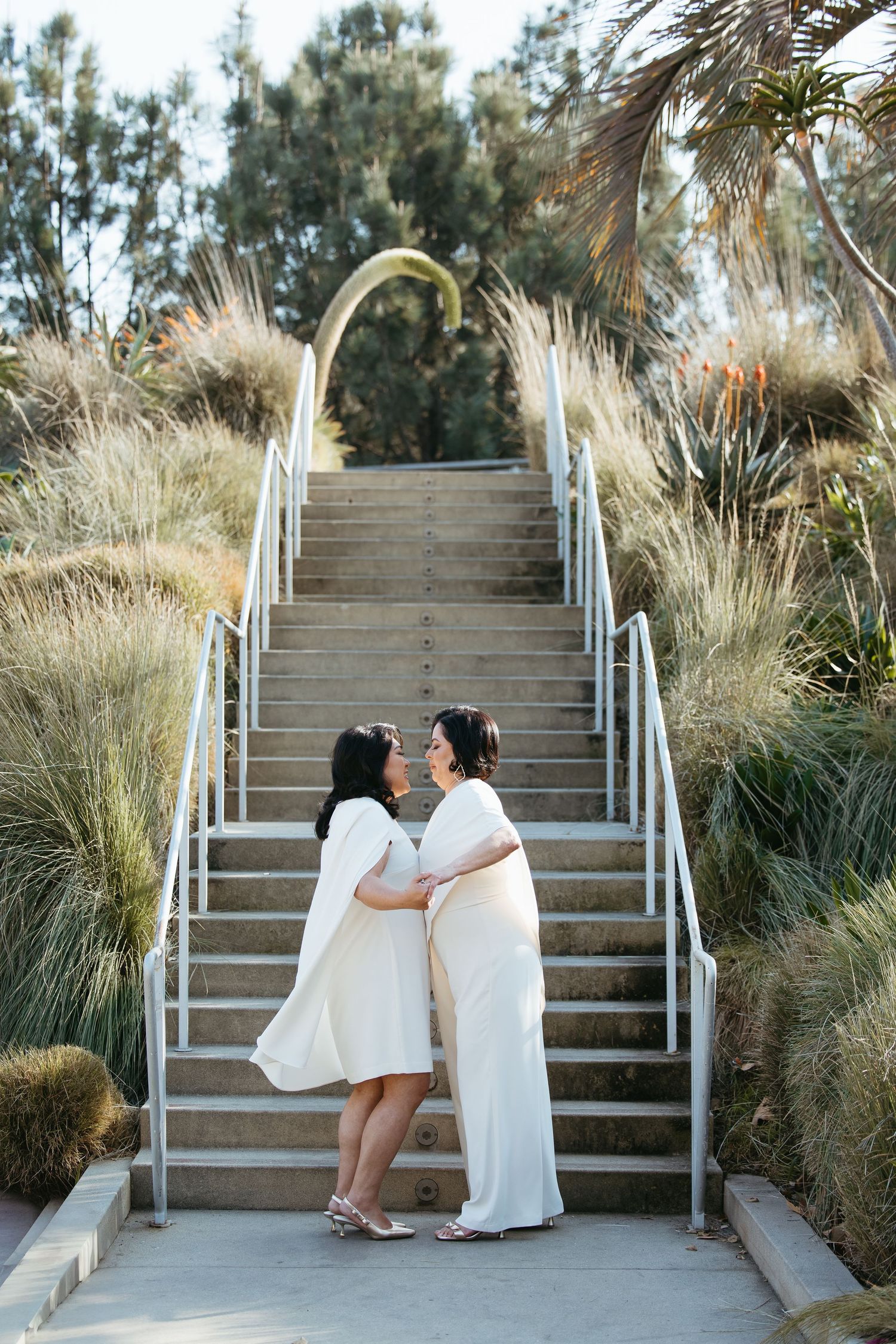 Two women in white dresses share a romantic moment at the base of outdoor stairs surrounded by lush greenery.