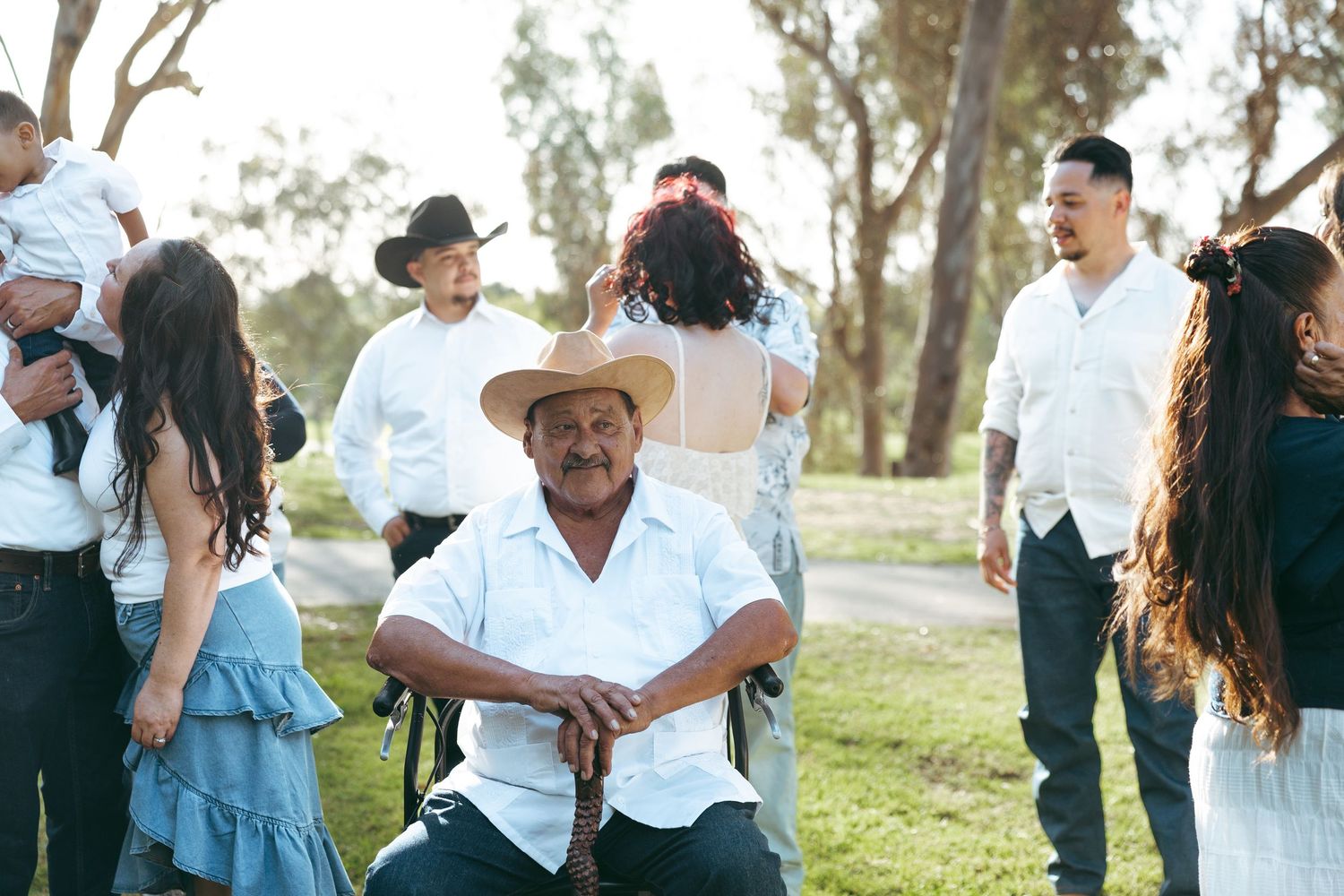 A smiling grandfather in a straw hat sits outdoors at a family gathering with others in white and blue attire among trees.