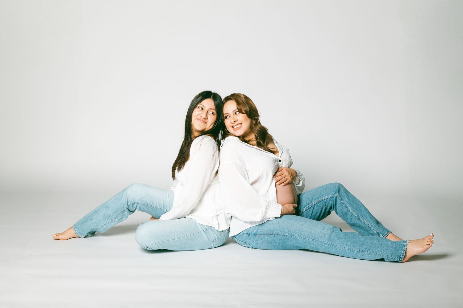 Two women in white sweaters and jeans sit back-to-back smiling on a white studio floor.