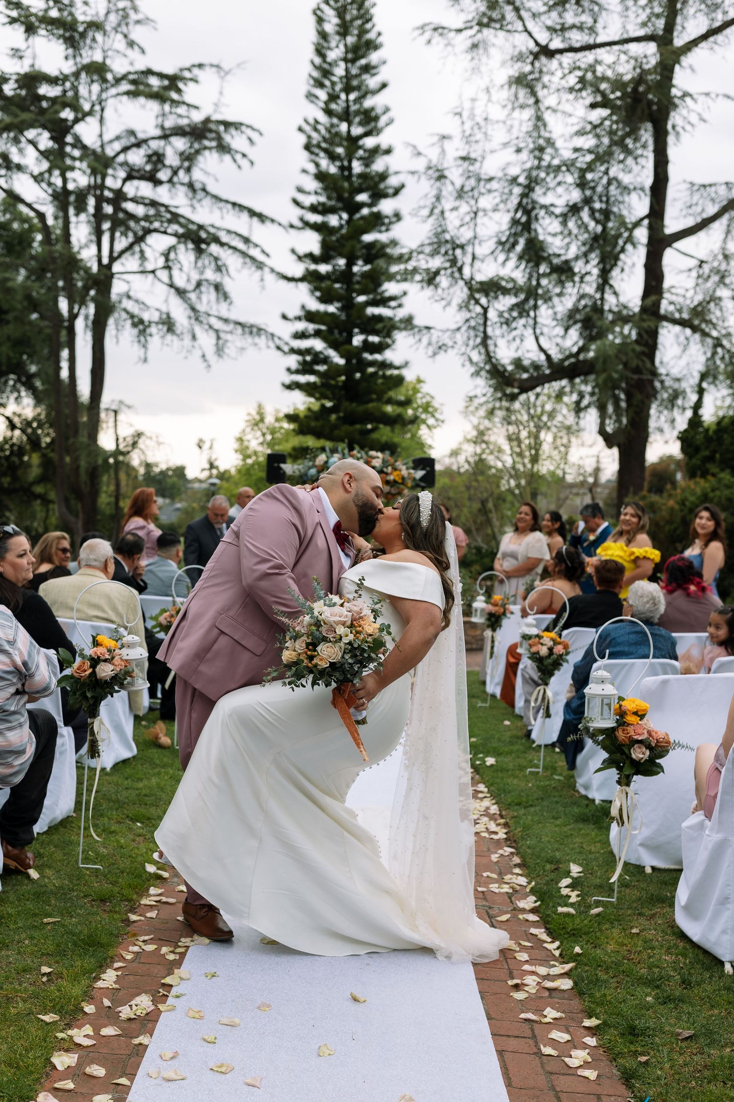 Bride and groom share a romantic kiss at outdoor wedding ceremony aisle surrounded by guests and tall trees.