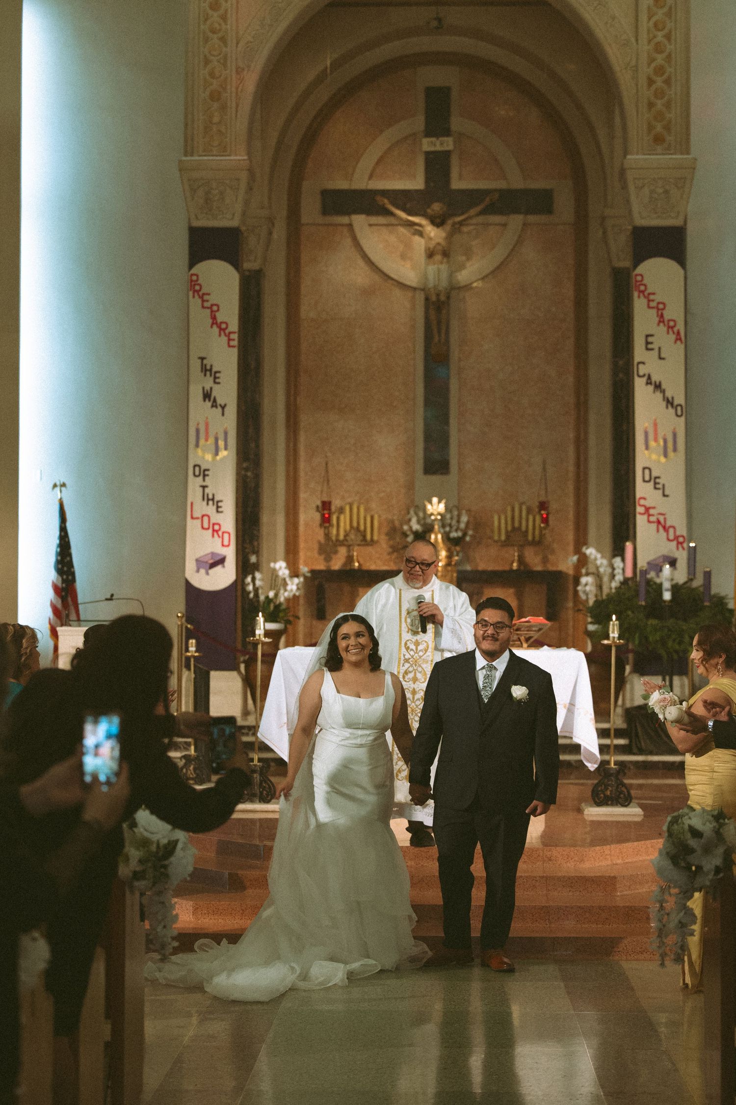 Newlyweds walk down Catholic church aisle after ceremony, bride in white gown, groom in suit, crucifix altar behind.