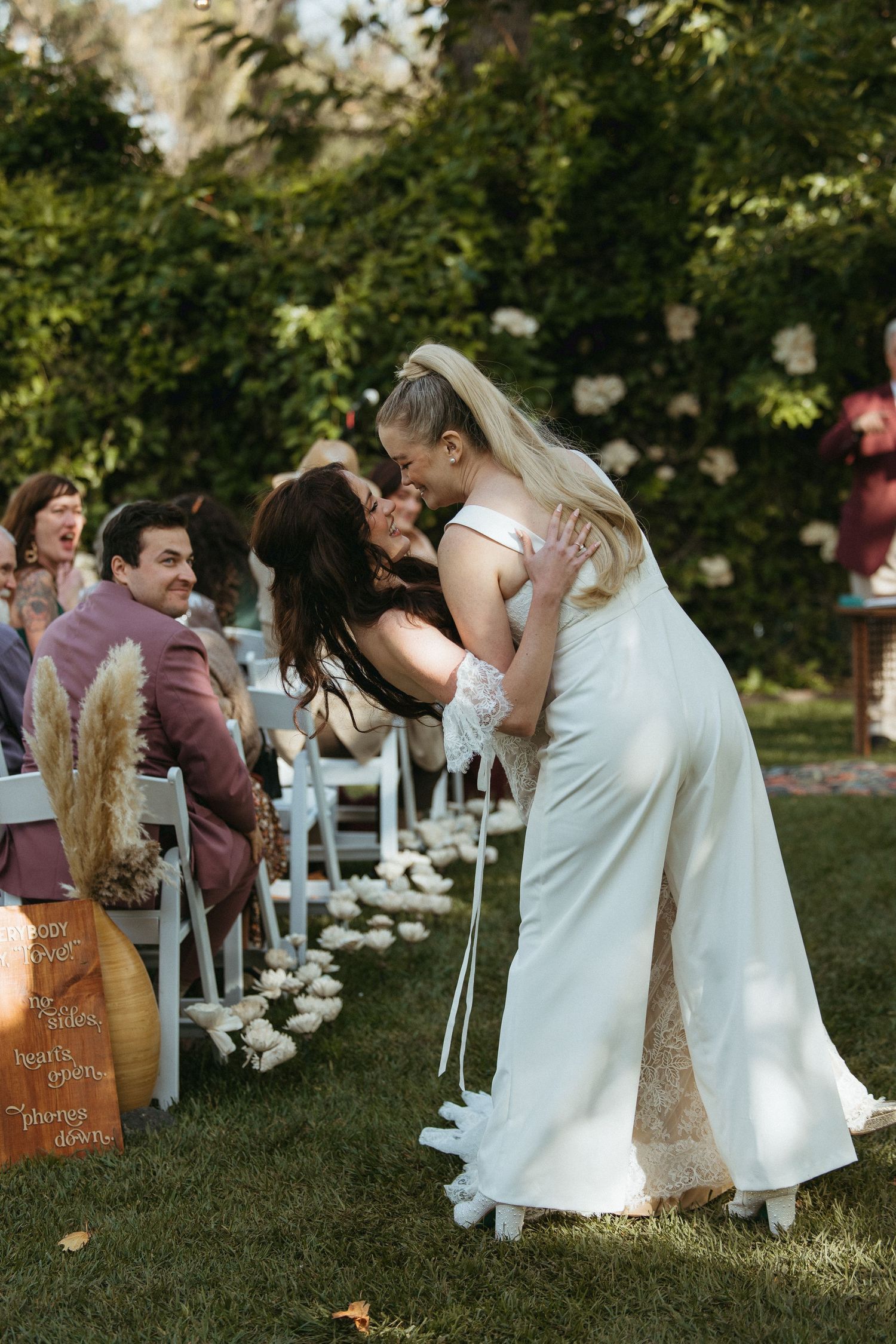 Two brides share a first kiss at an outdoor garden wedding ceremony while guests watch from white chairs.