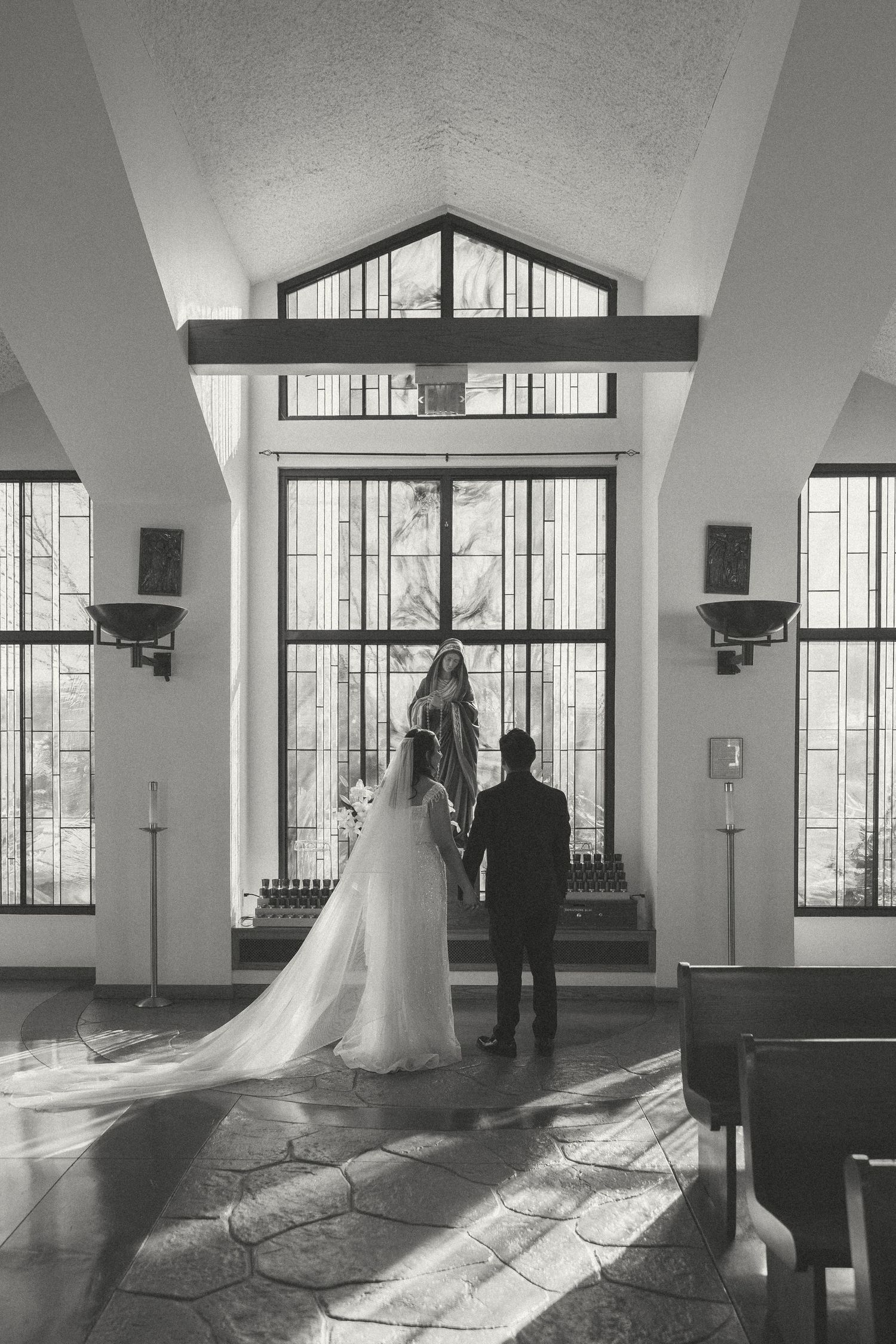 Black and white photo of bride and groom holding hands before a stained glass chapel window.