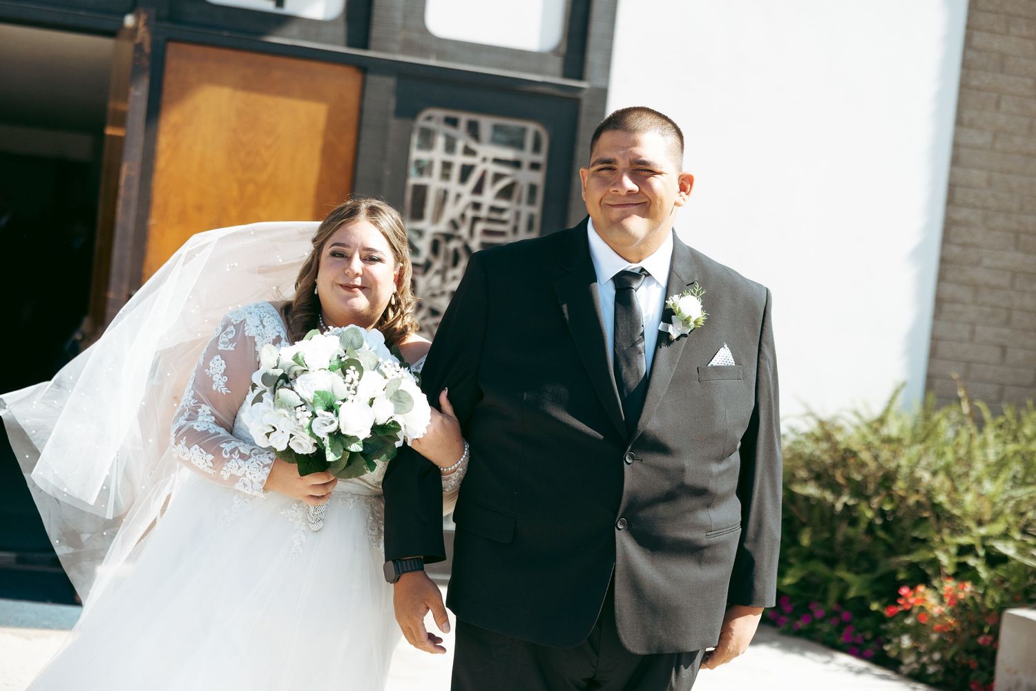 Bride in white lace gown holding bouquet walks with groom in gray suit outside church on wedding day.