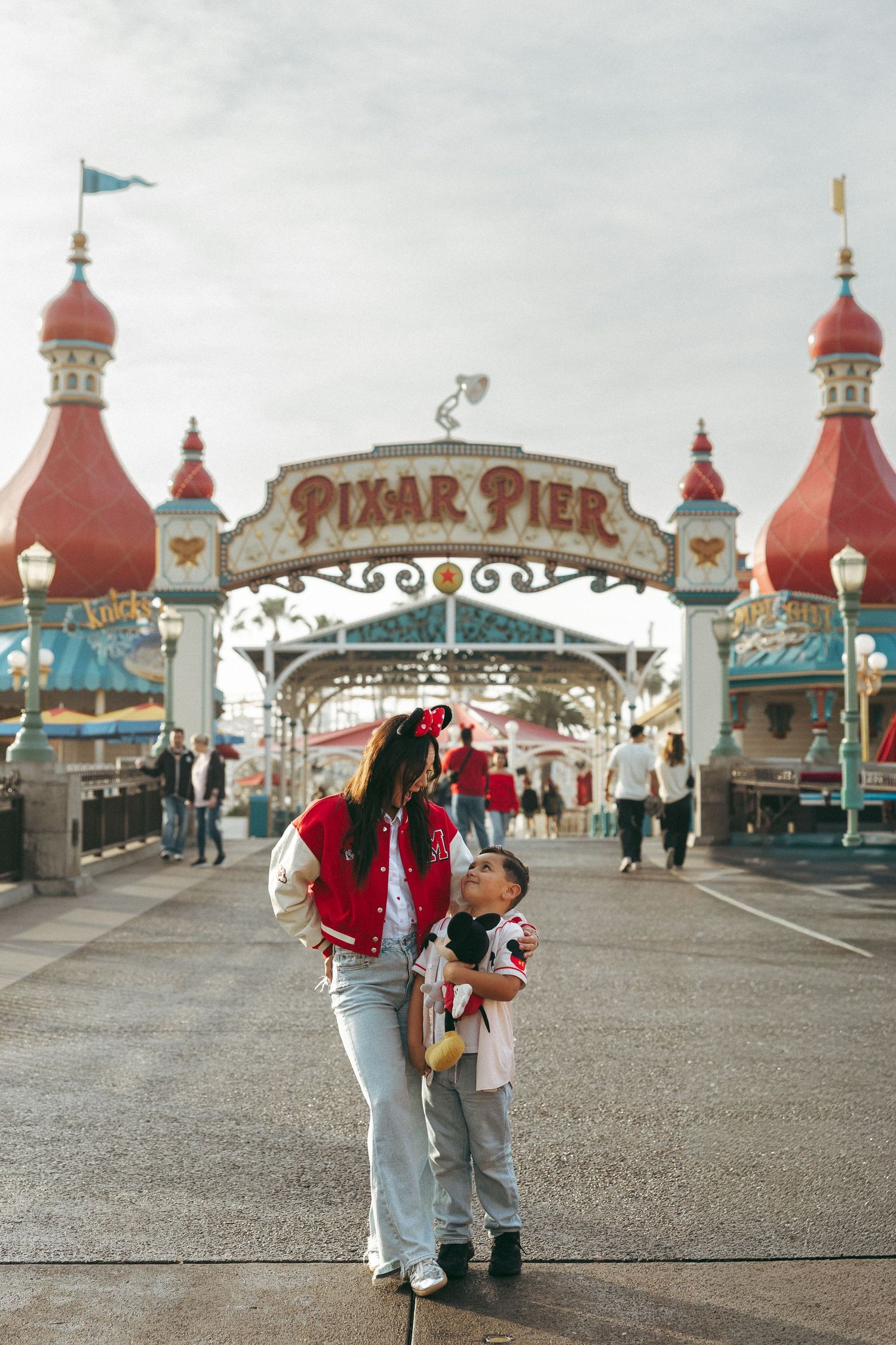 A mother and child pose at Pixar Pier entrance at Disney California Adventure Park, child holding Mickey Mouse plush.