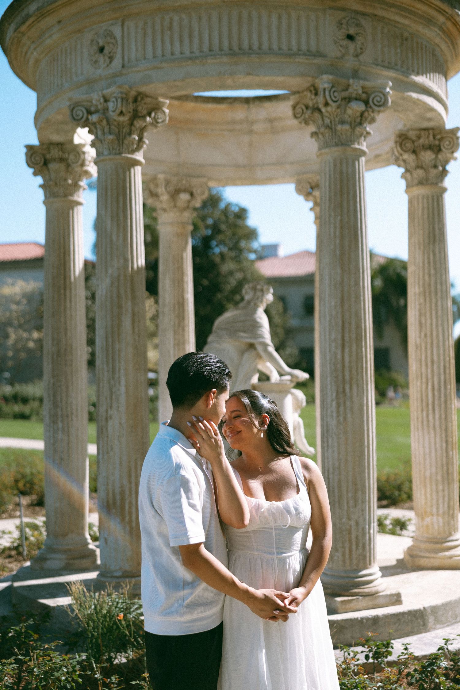 Couple embracing near classical marble columns and statue in a sunlit garden, woman in white dress. Garden engagement session.