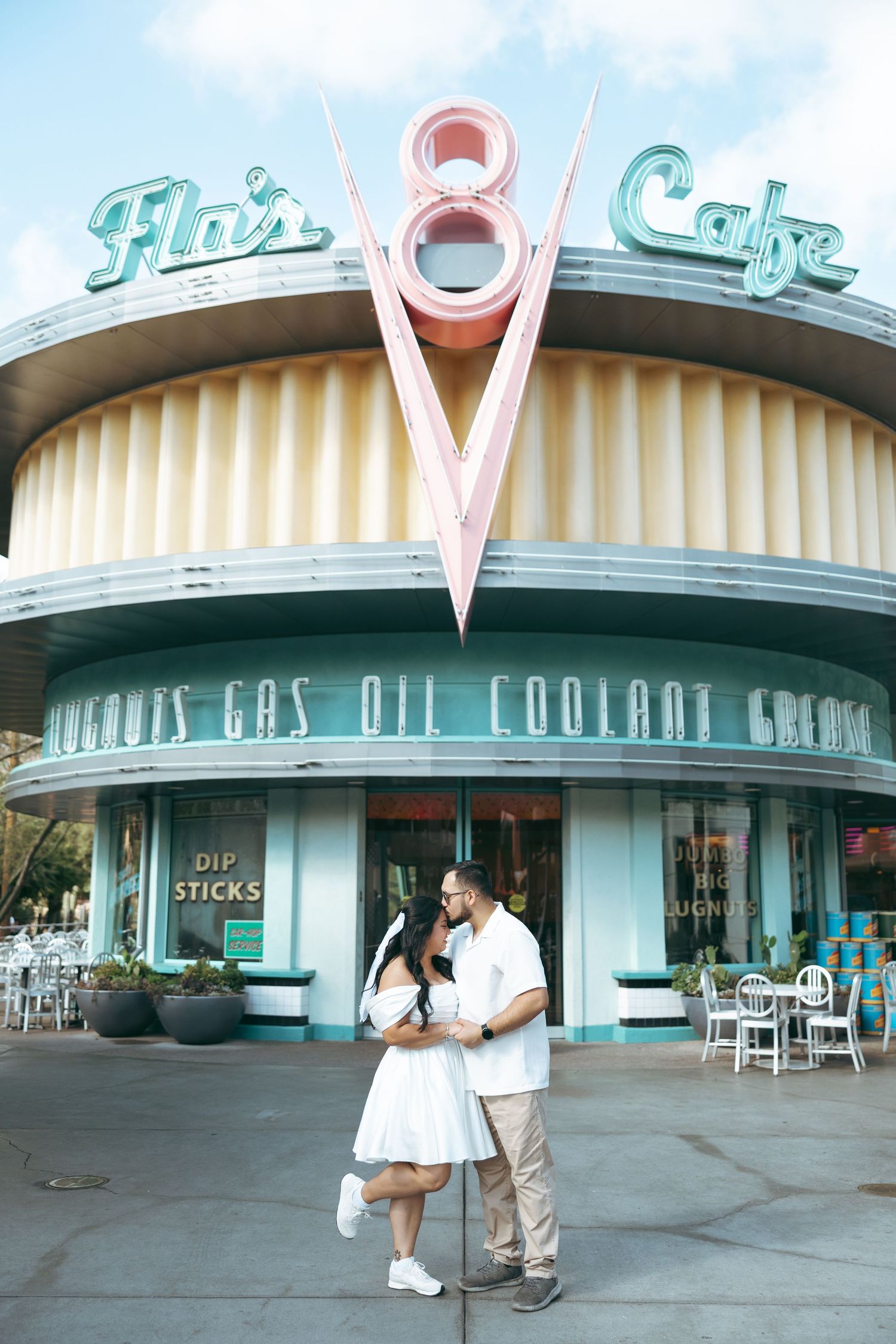 Couple sharing a romantic kiss in front of Flo's V8 Cafe at Disney's Cars Land, woman in white dress.
