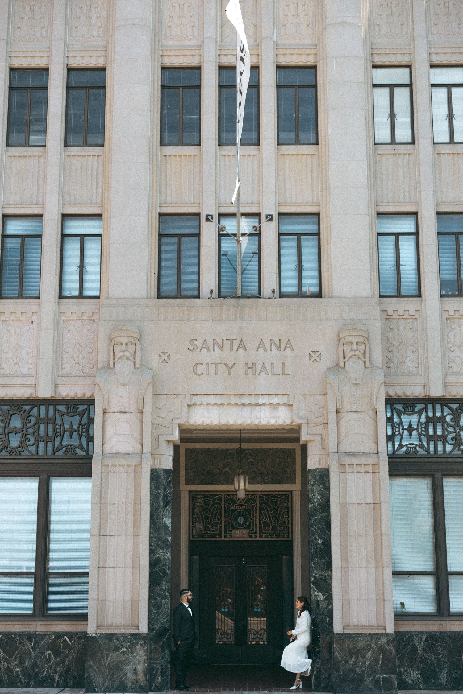 Engagement session where couple poses at Santa Ana City Hall entrance, featuring Art Deco architecture with ornate doors and stone facade.