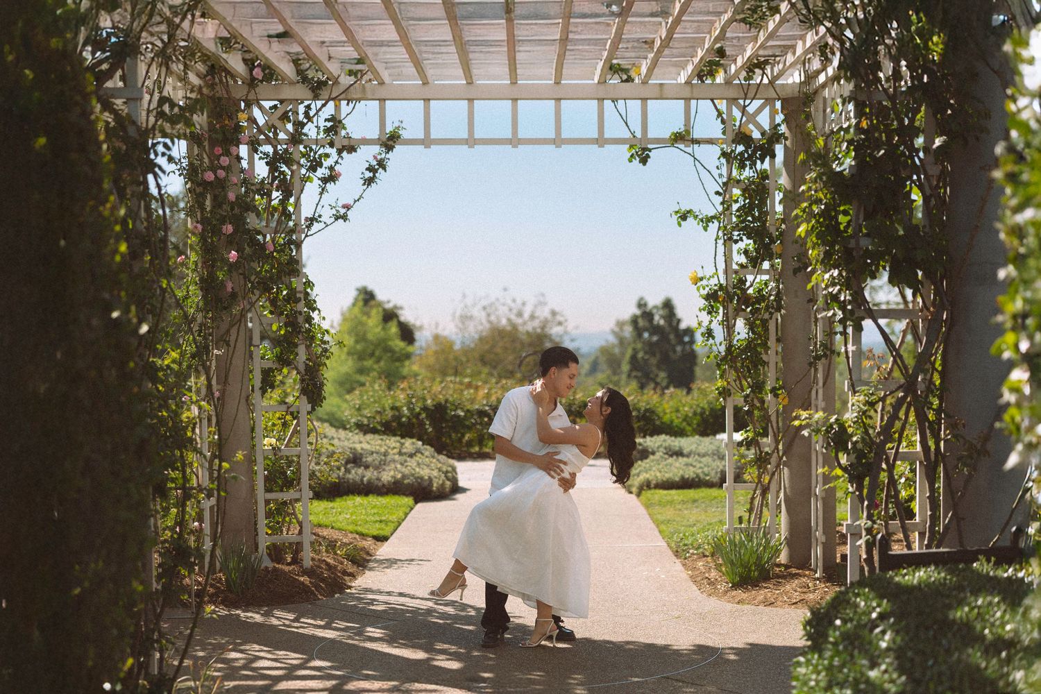 Bride and groom sharing a romantic dip kiss under a vine-covered pergola in a sunny garden.