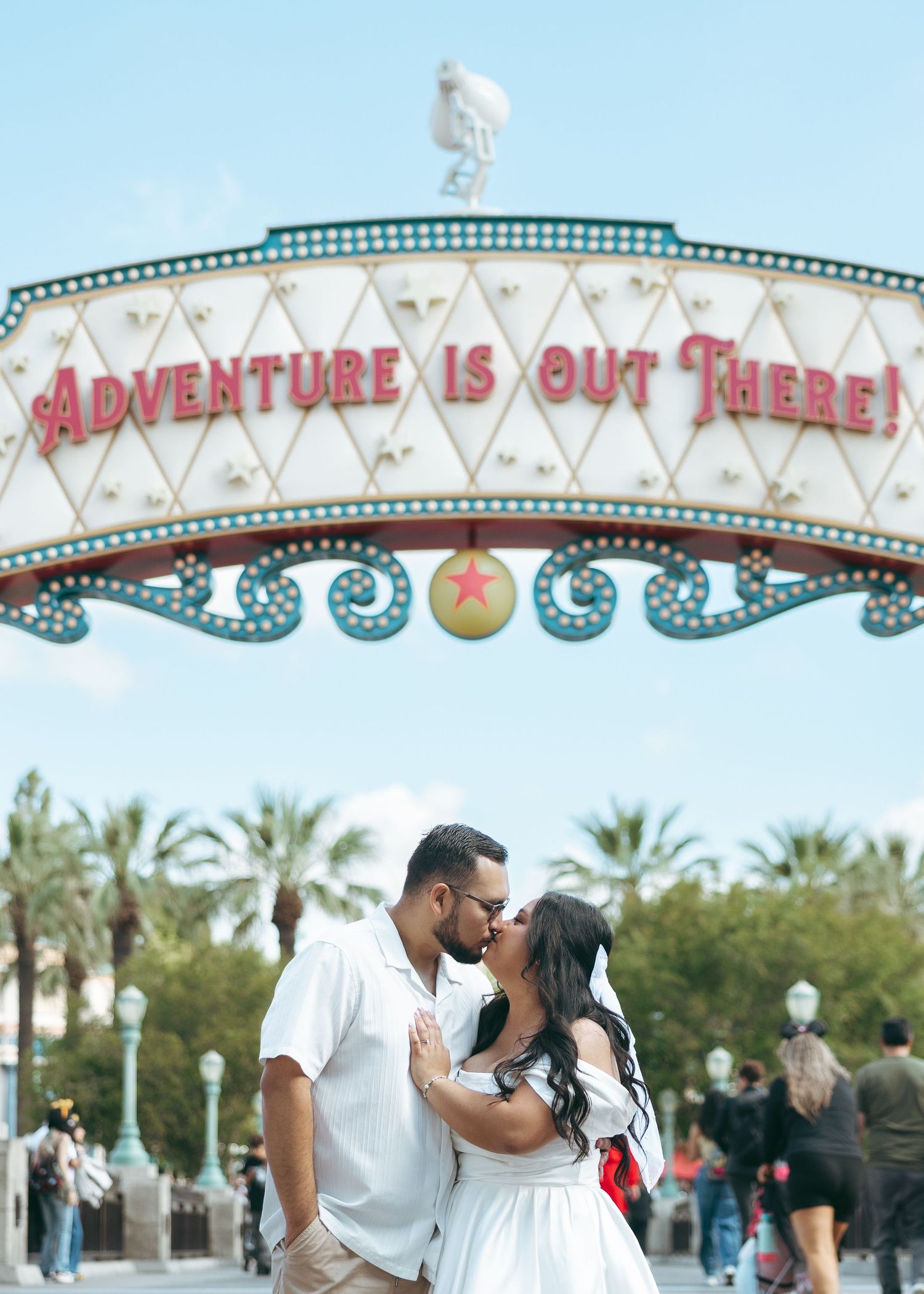 Couple kissing under Adventure Is Out There sign at Disney California Adventure park with palm trees in background.