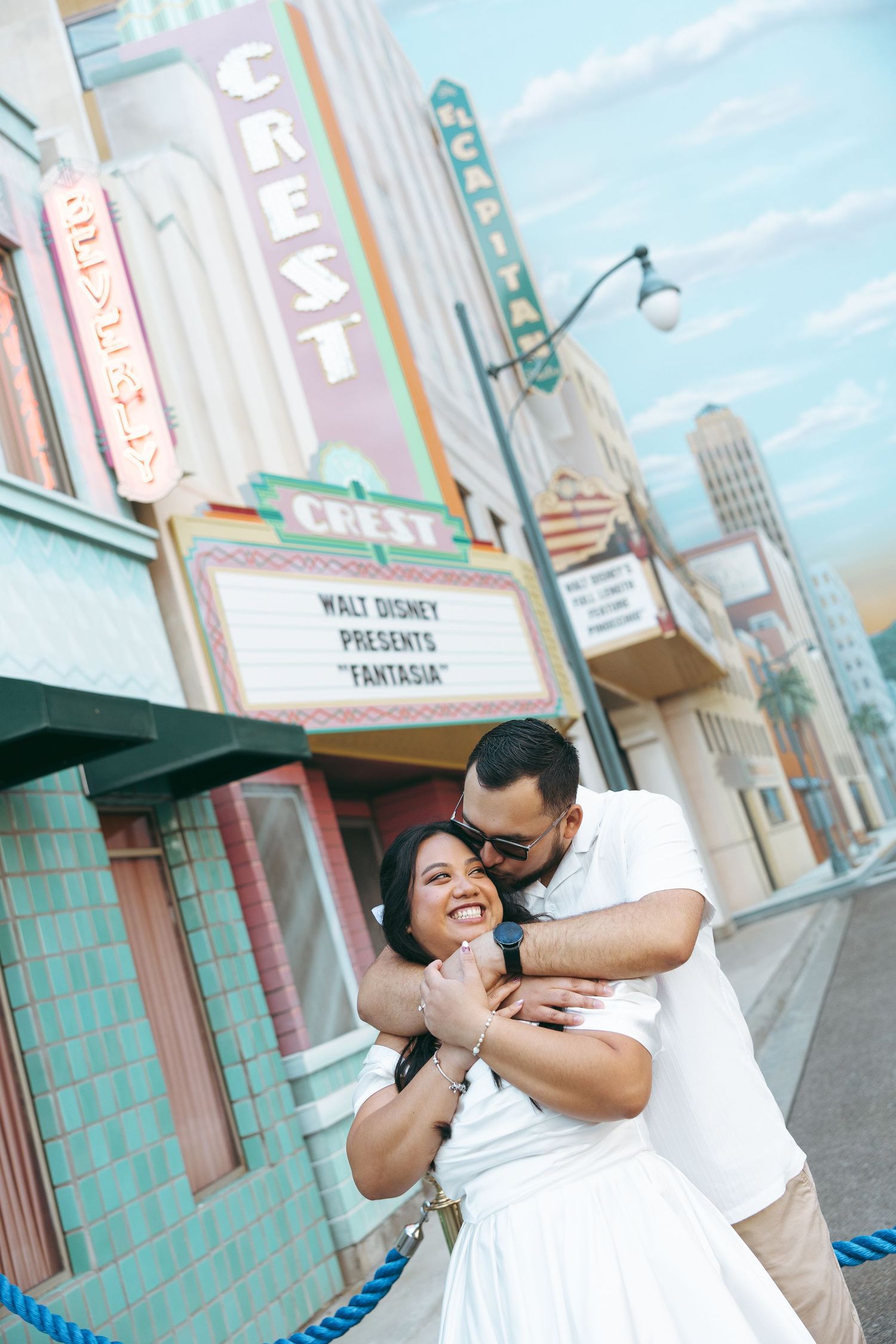 Couple embracing and smiling in front of the Crest Theatre at Disney, wearing matching white outfits.