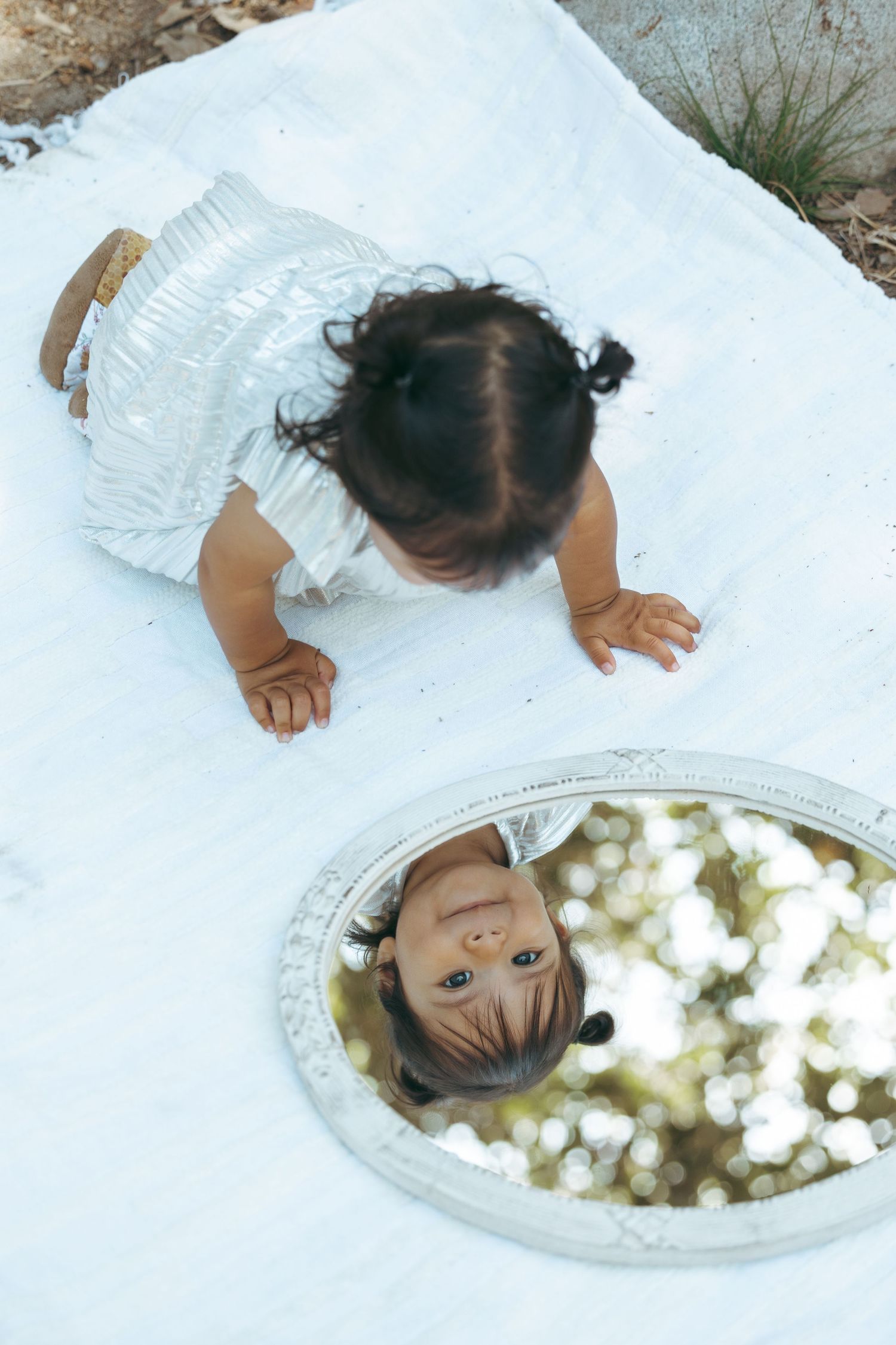 Baby in white dress crawling on white blanket, looking at reflection in oval mirror outdoors.