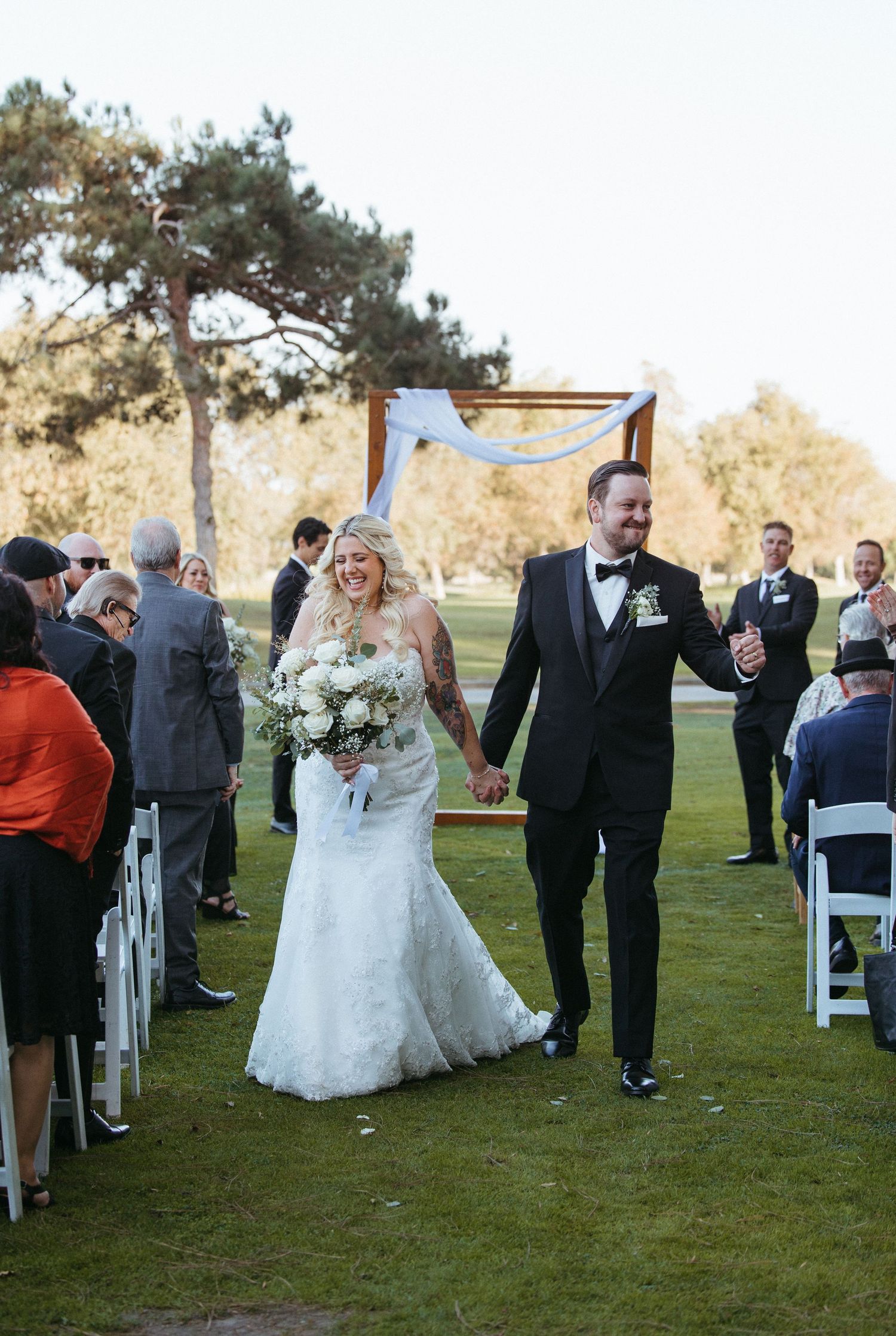 Happy newlyweds walk down outdoor wedding aisle, bride in lace mermaid gown holding white bouquet, groom in black tuxedo.