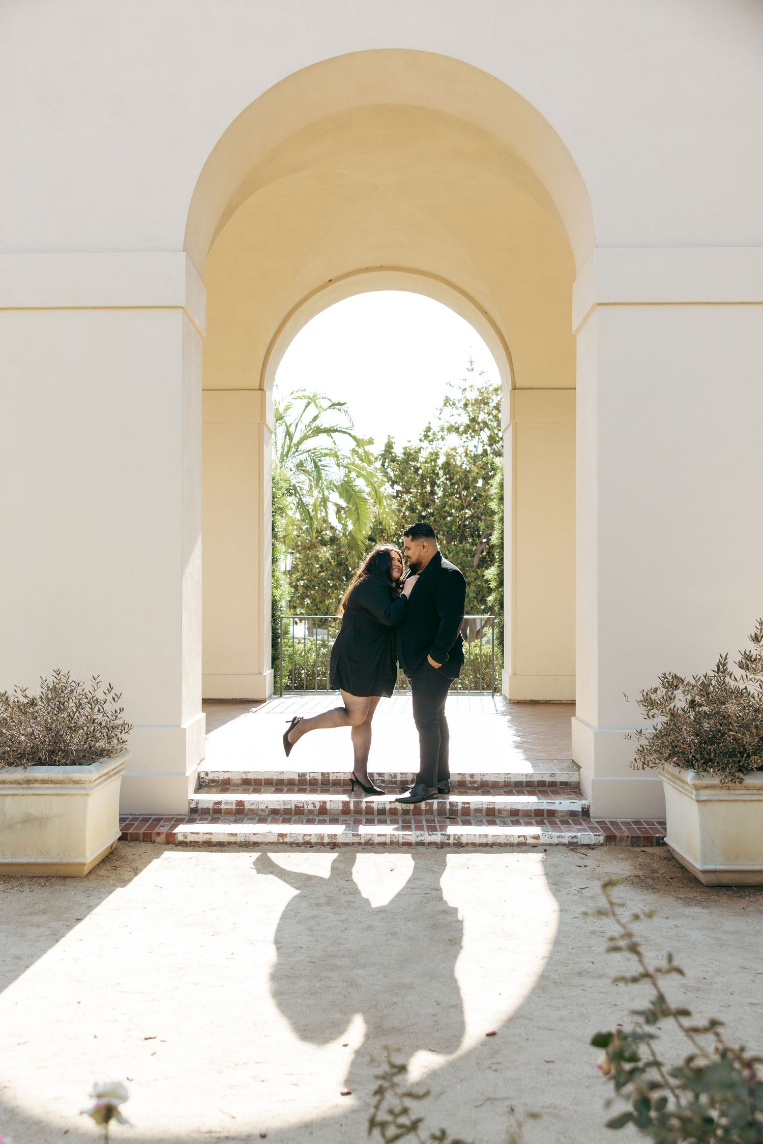 Couple sharing a romantic kiss under an elegant yellow arch, casting shadows on a sunlit courtyard with potted plants. Pasadena City Hall.