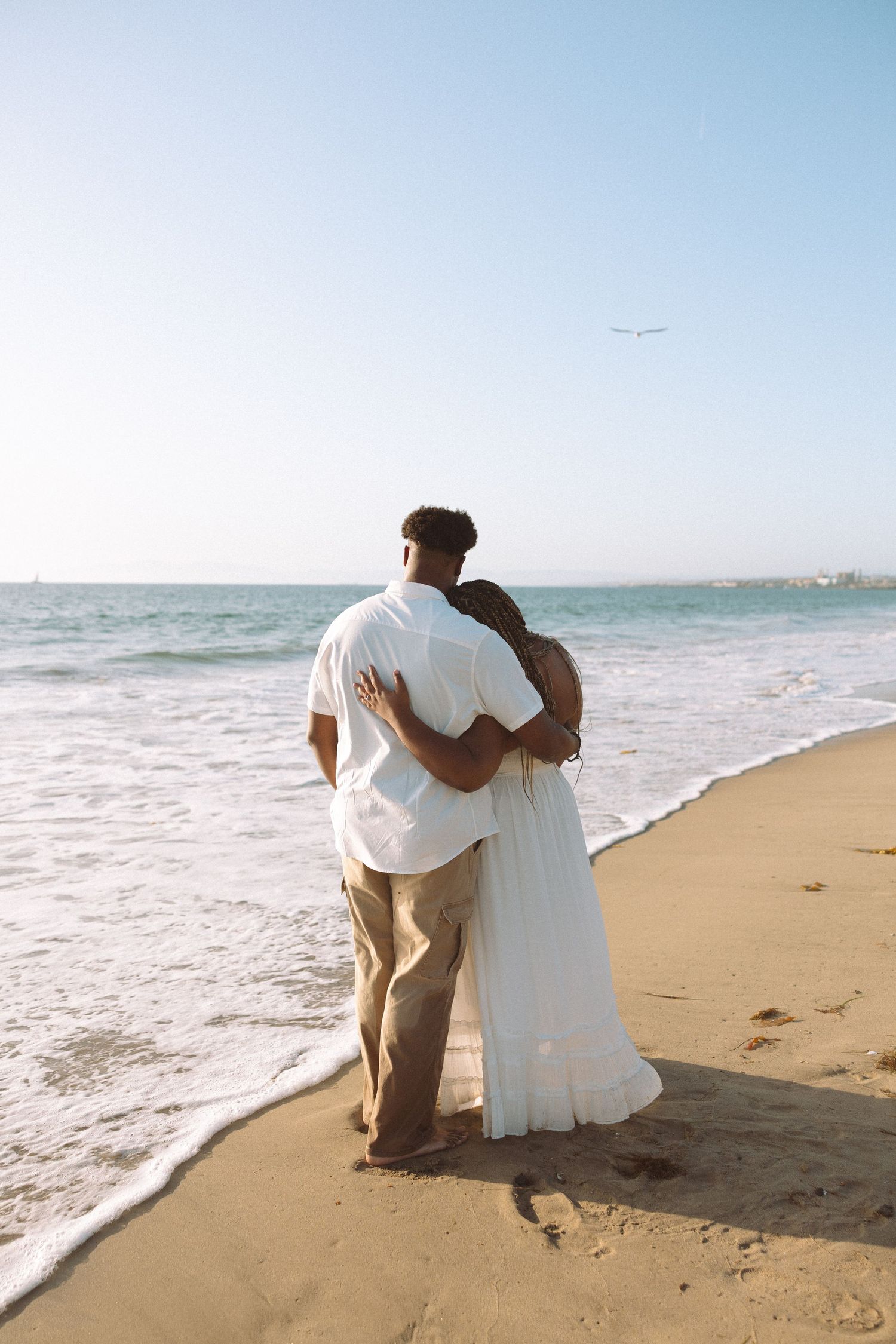 Couple embracing on a sandy beach, gazing at ocean waves under a clear blue sky. Engagement Beach session in the South Bay Area.