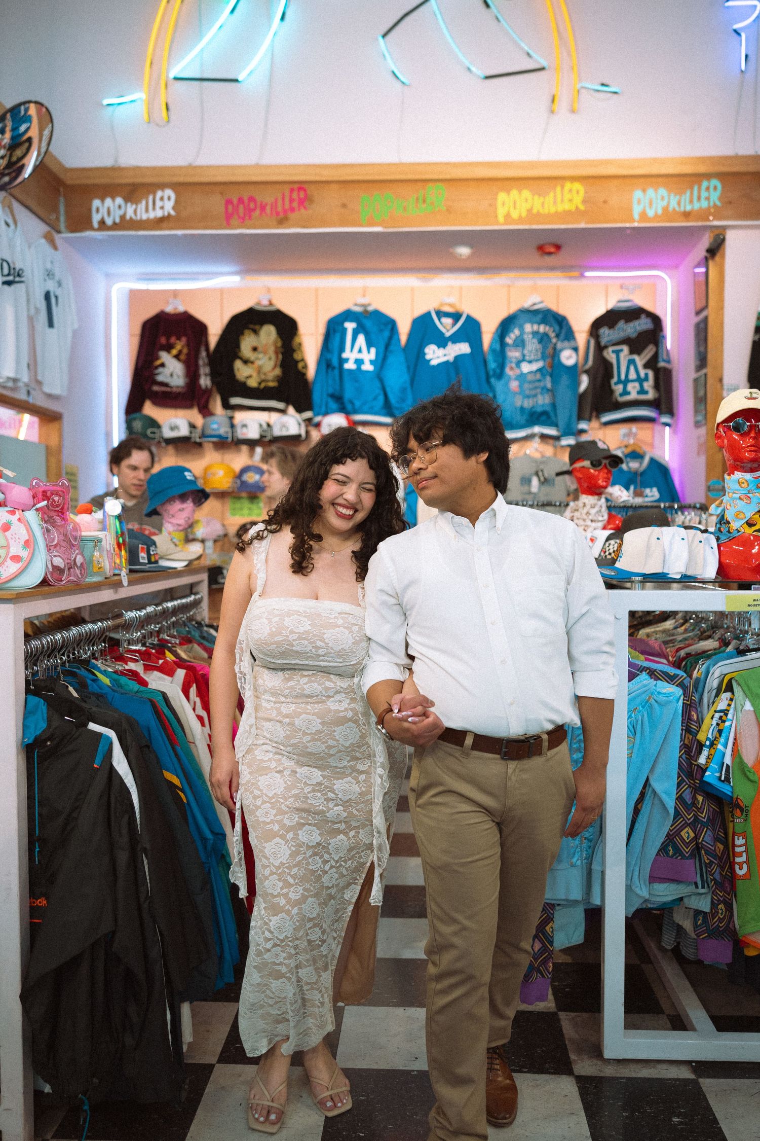 Couple holds hands and smiles at each other inside a colorful vintage clothing store with neon signs.