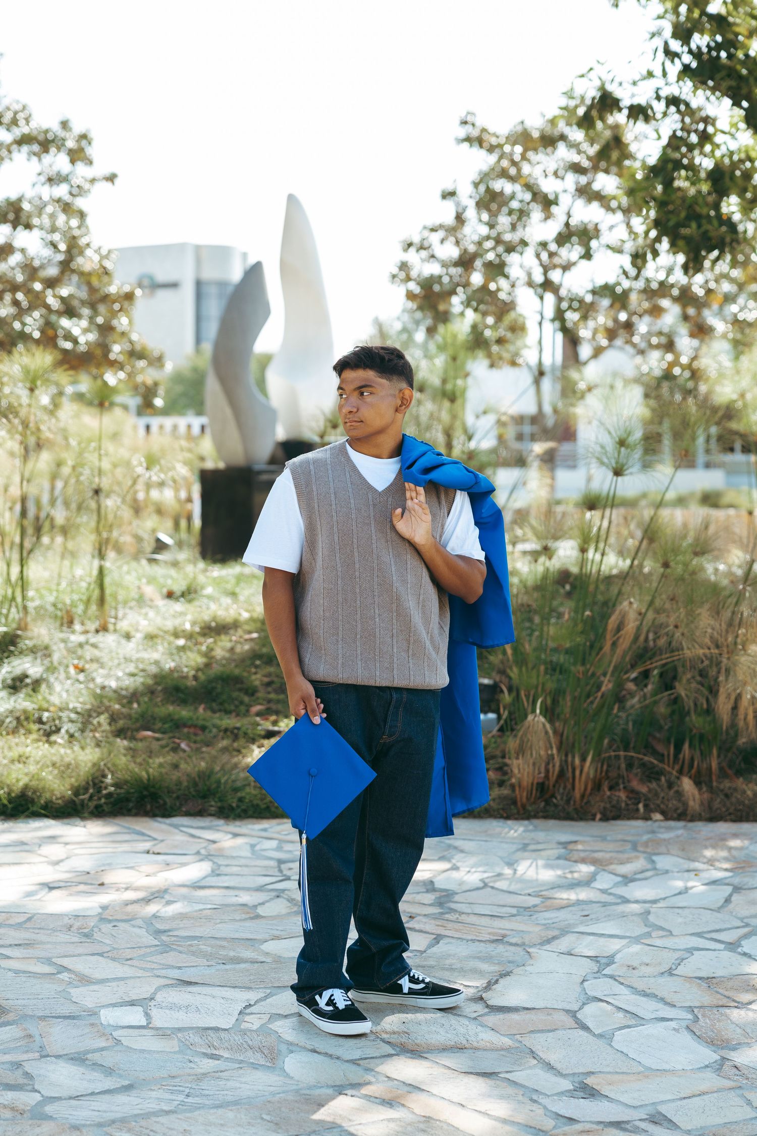 Young man holding blue graduation cap and gown outdoors on stone path with trees and sculpture in background.