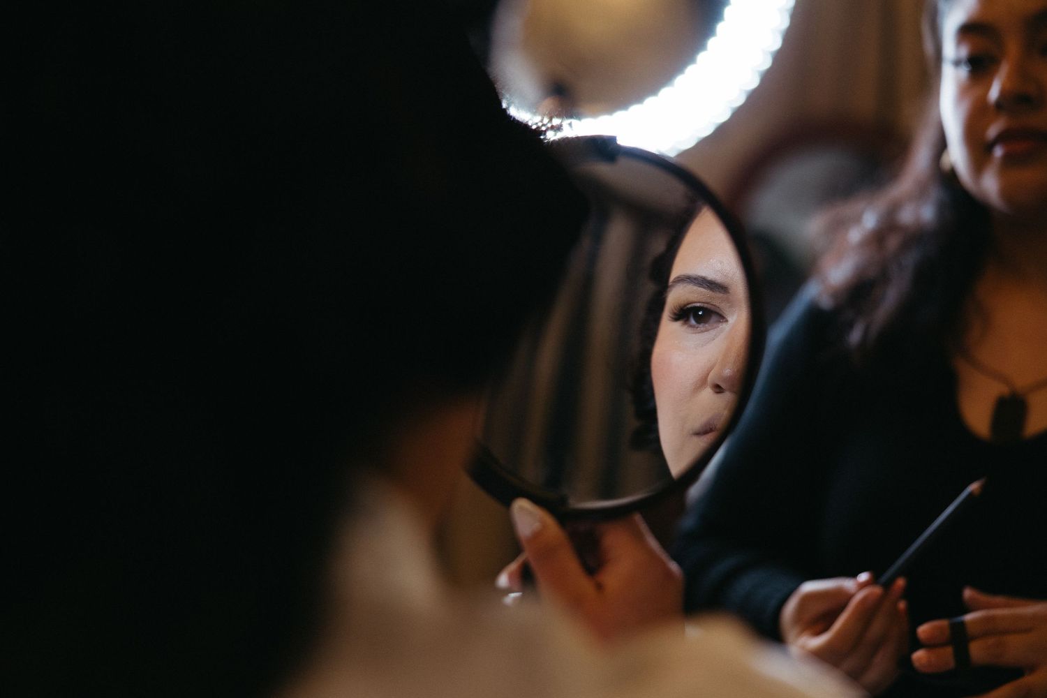 Woman getting makeup done, reflected in a round mirror with ring light in a dimly lit studio.