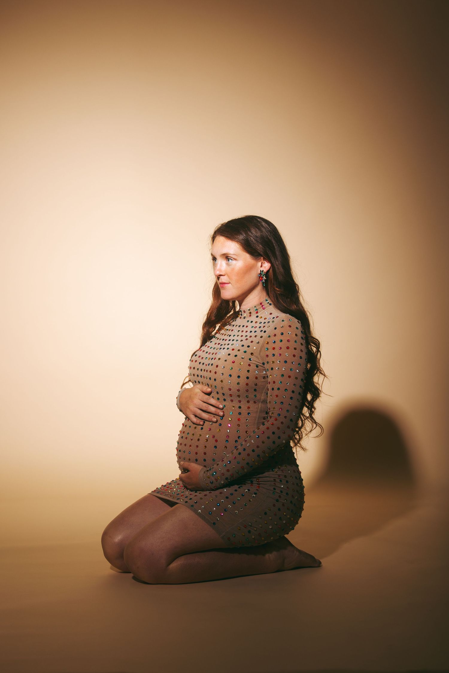 Pregnant woman in beaded dress kneeling on floor, cradling baby bump in studio portrait.