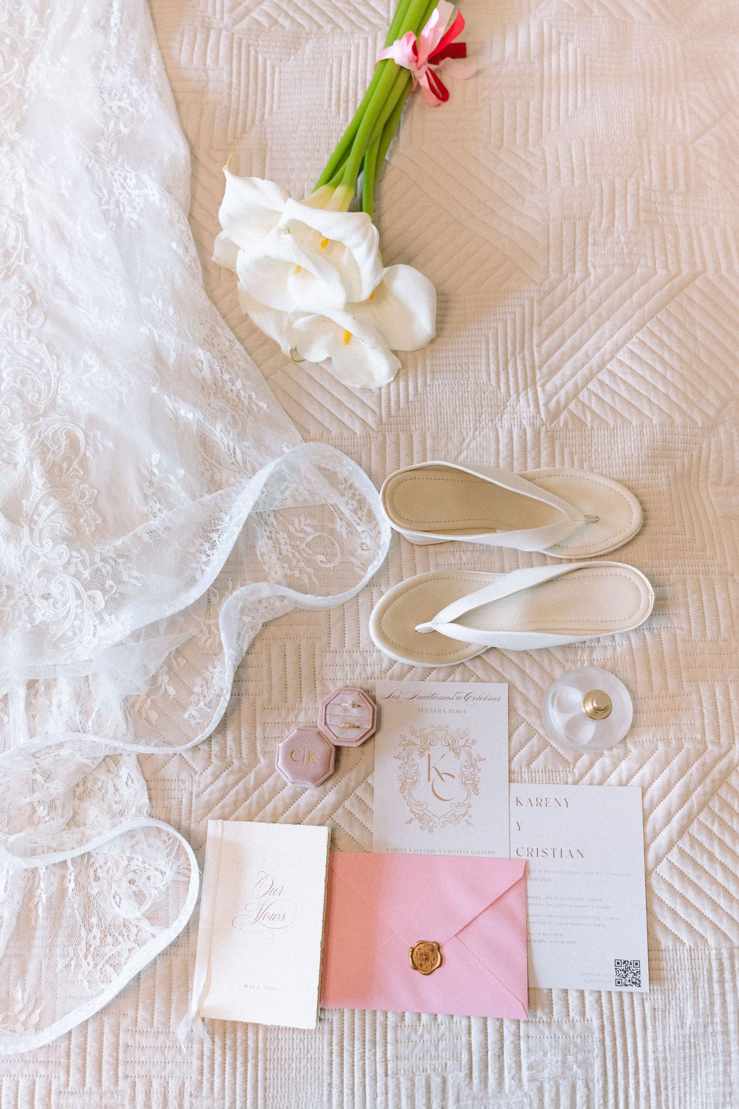 Wedding flat lay featuring lace bridal gown, white sandals, floral bouquet, pink envelope, and elegant invitation suite.
