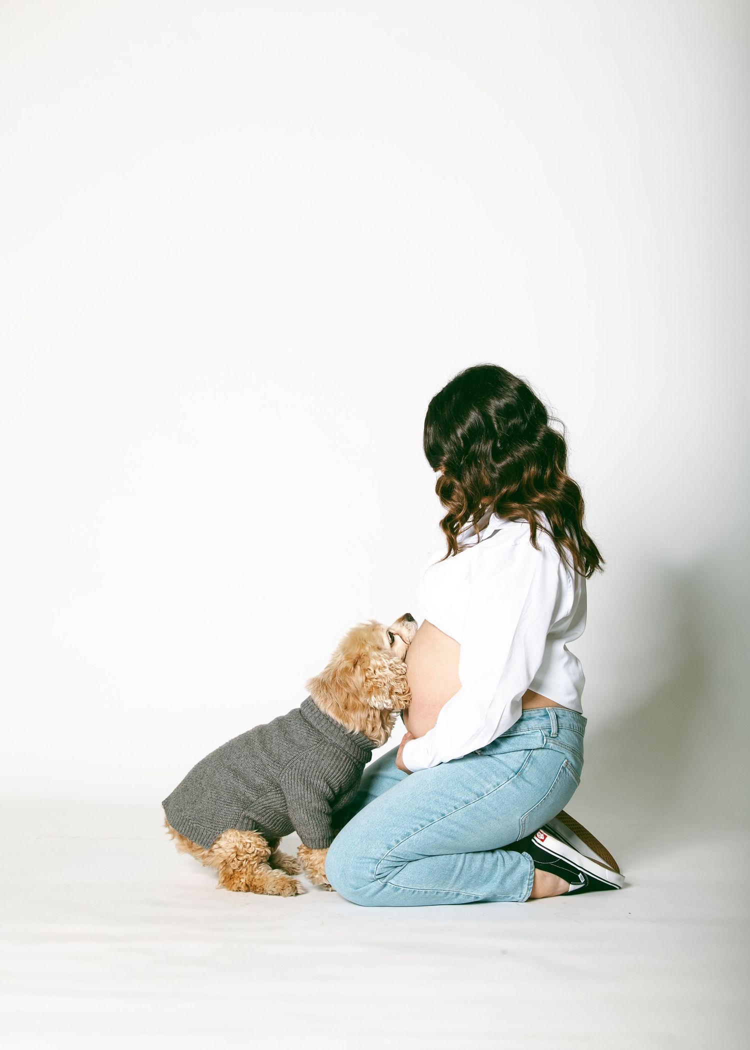 A brunette woman sits on the floor bonding with her small curly-haired dog wearing a gray sweater.