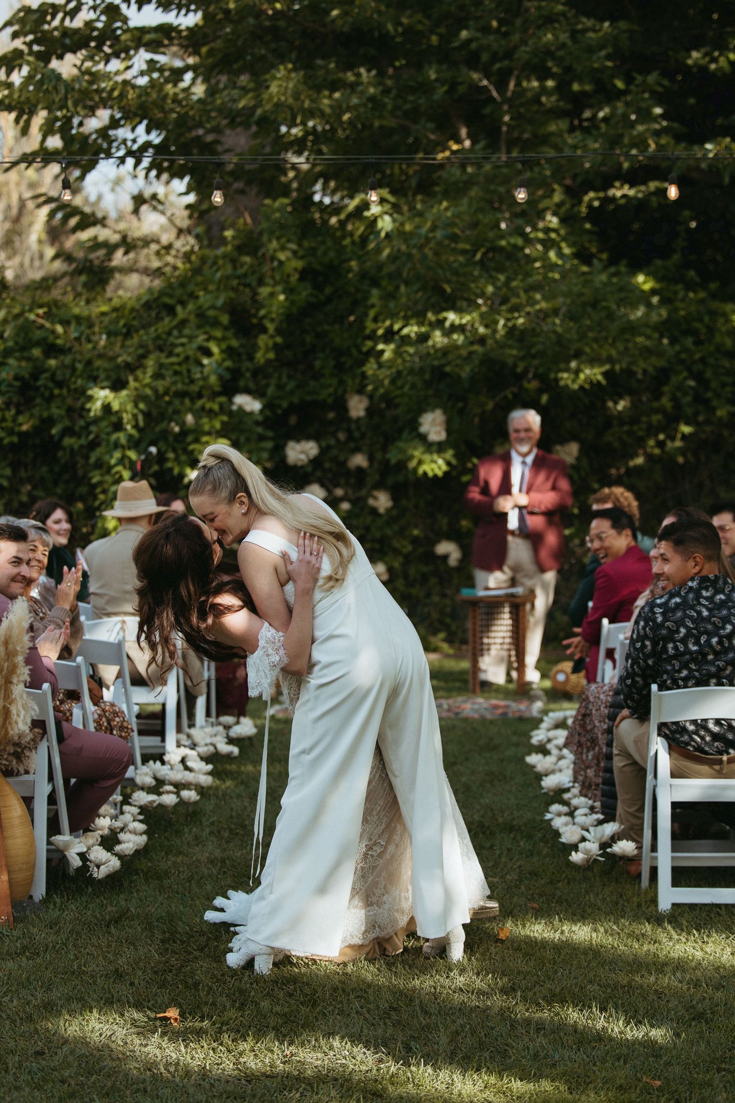 Two brides share a romantic dip kiss while walking down the aisle at an outdoor wedding ceremony.