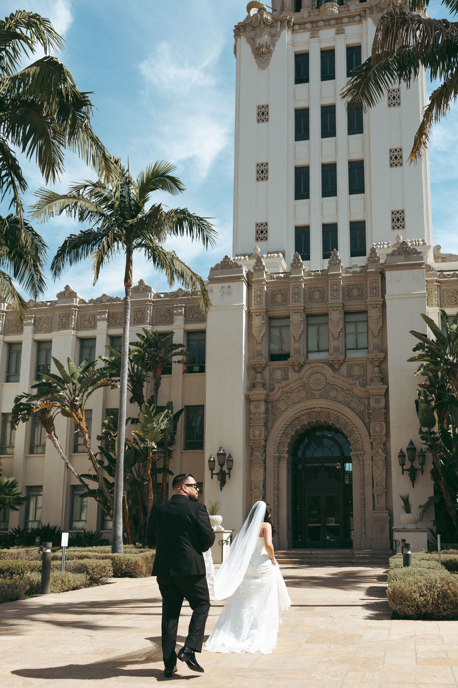 Bride and groom posing in front of an ornate building with palm trees under a blue sky.