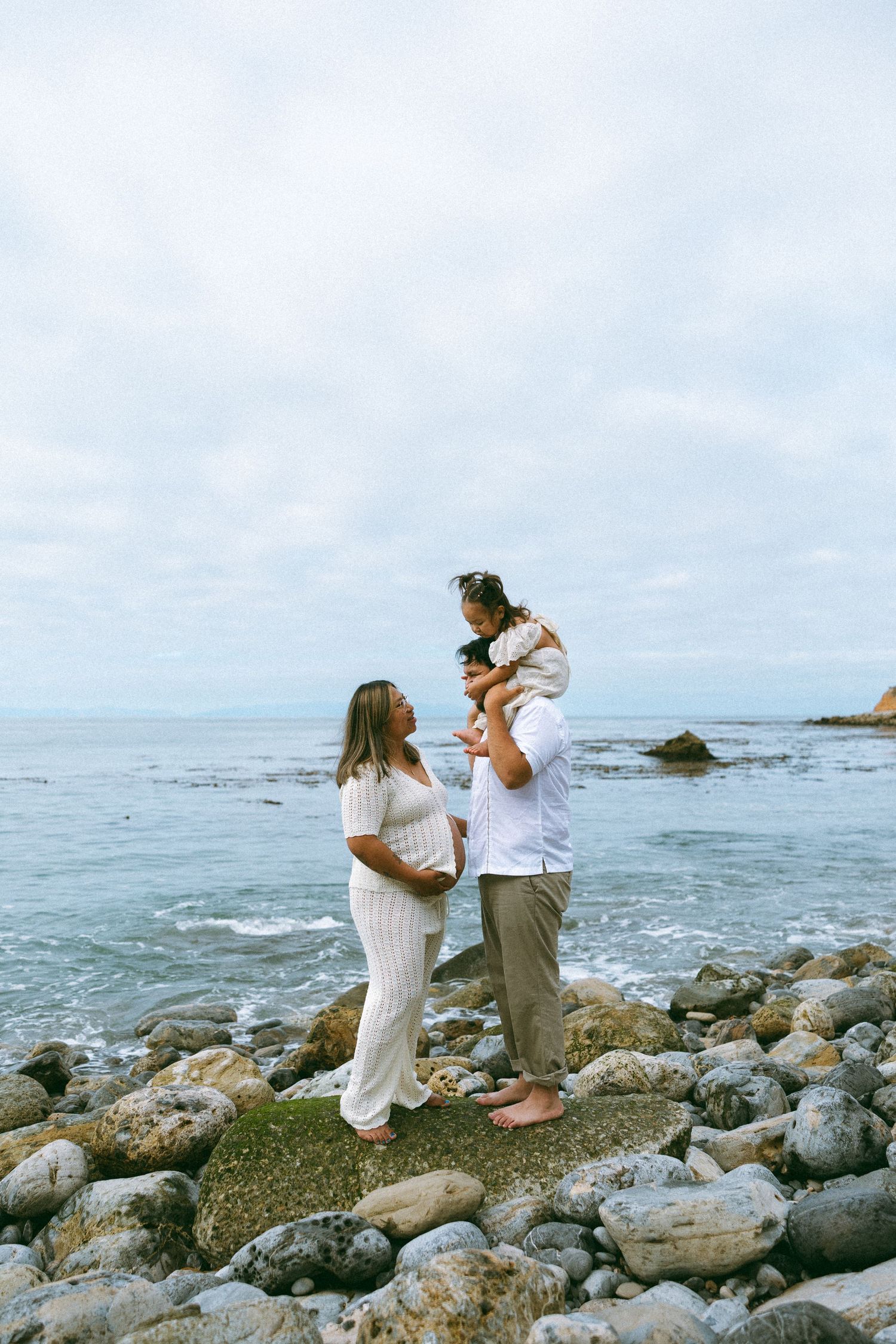 Pregnant woman and man holding child stand on rocky beach shore with ocean waves and cloudy sky behind them. Palos verdes maternity session.