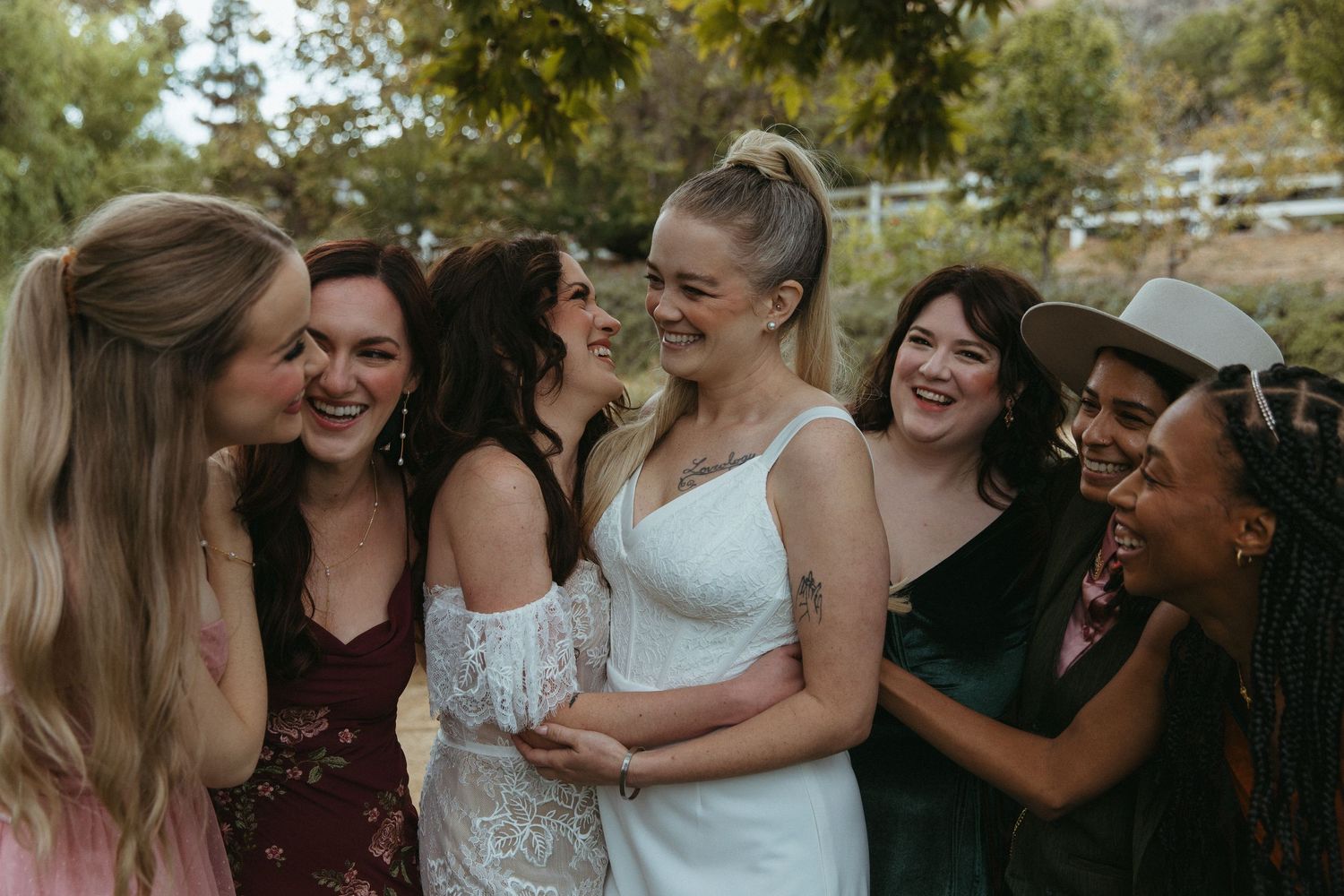 Brides in white dress and jumpsuit laughing with bridesmaids in colorful gowns outdoors at a wedding celebration. Madonna Inn wedding.