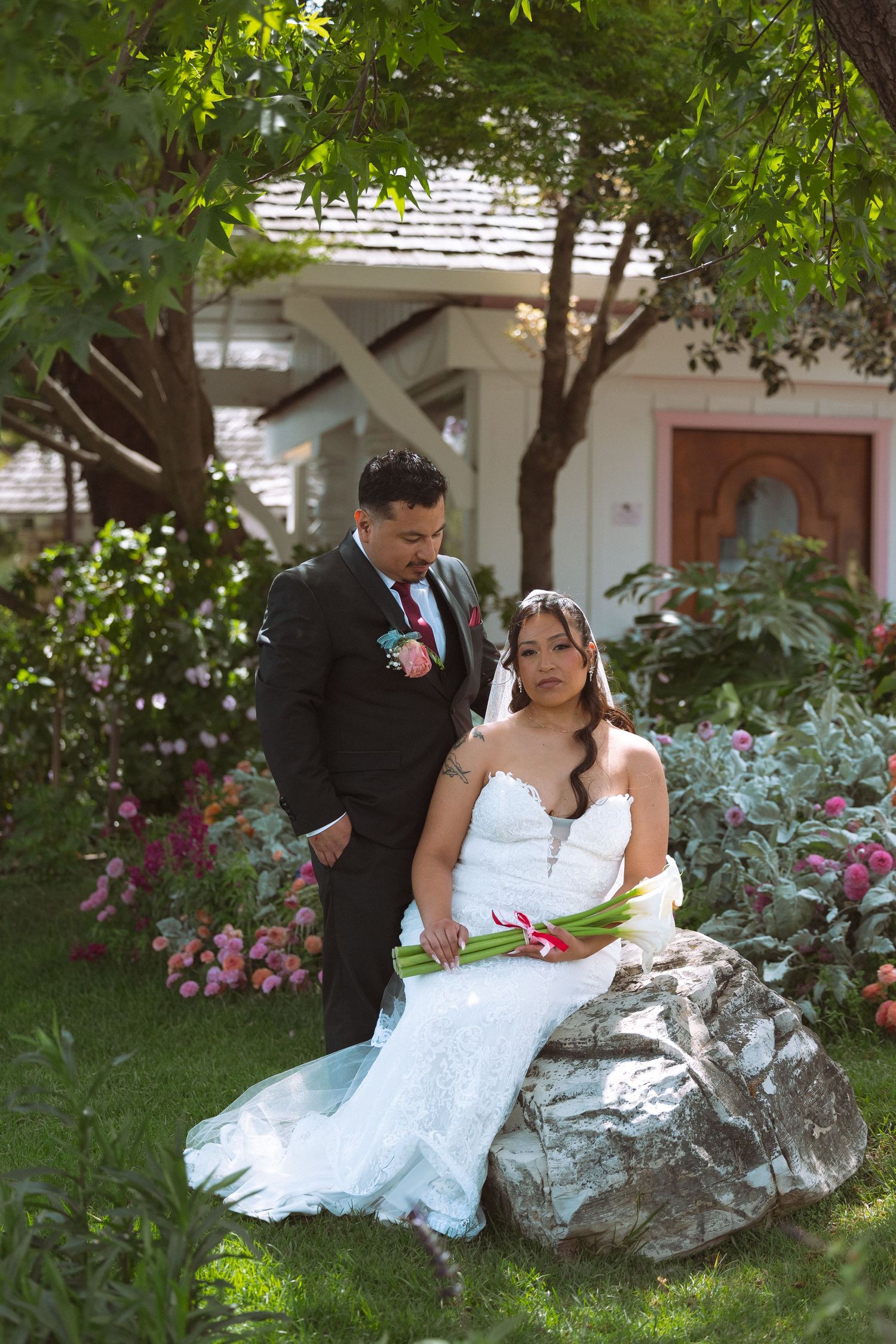 Bride in lace mermaid gown seated on a rock with groom in black suit in a lush garden setting.