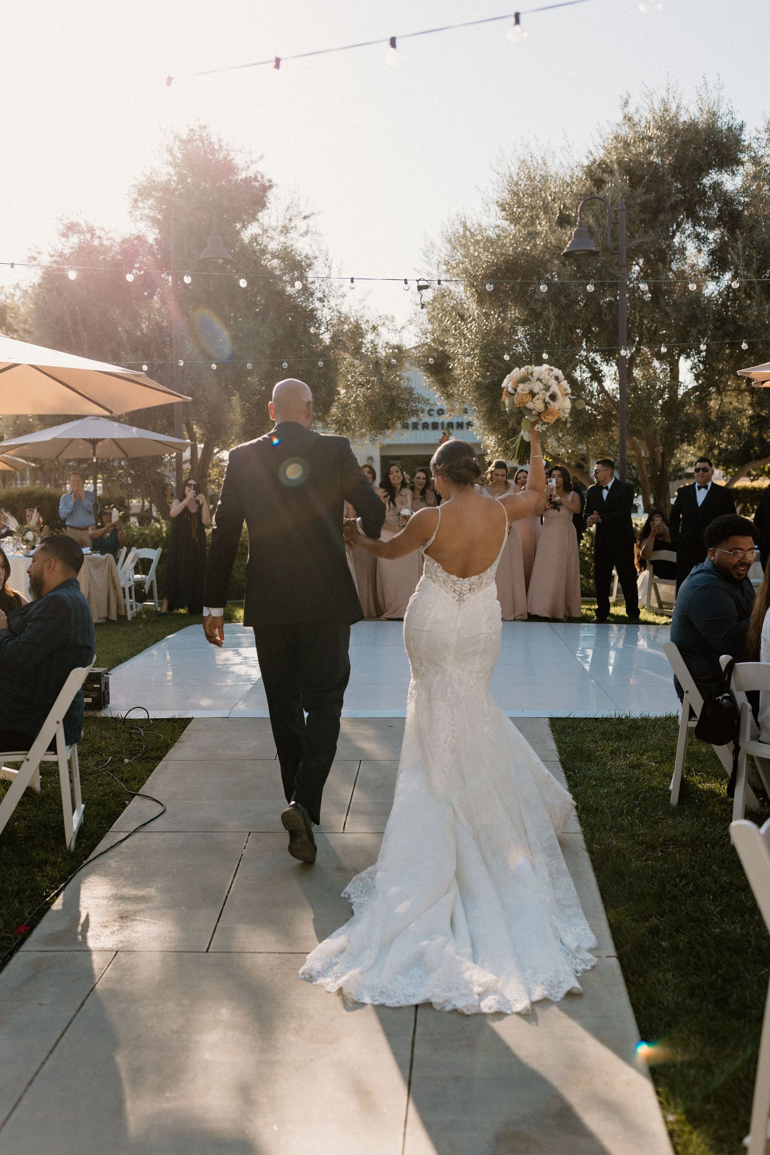 Bride raising bouquet while walking with groom down outdoor aisle at golden hour wedding reception.