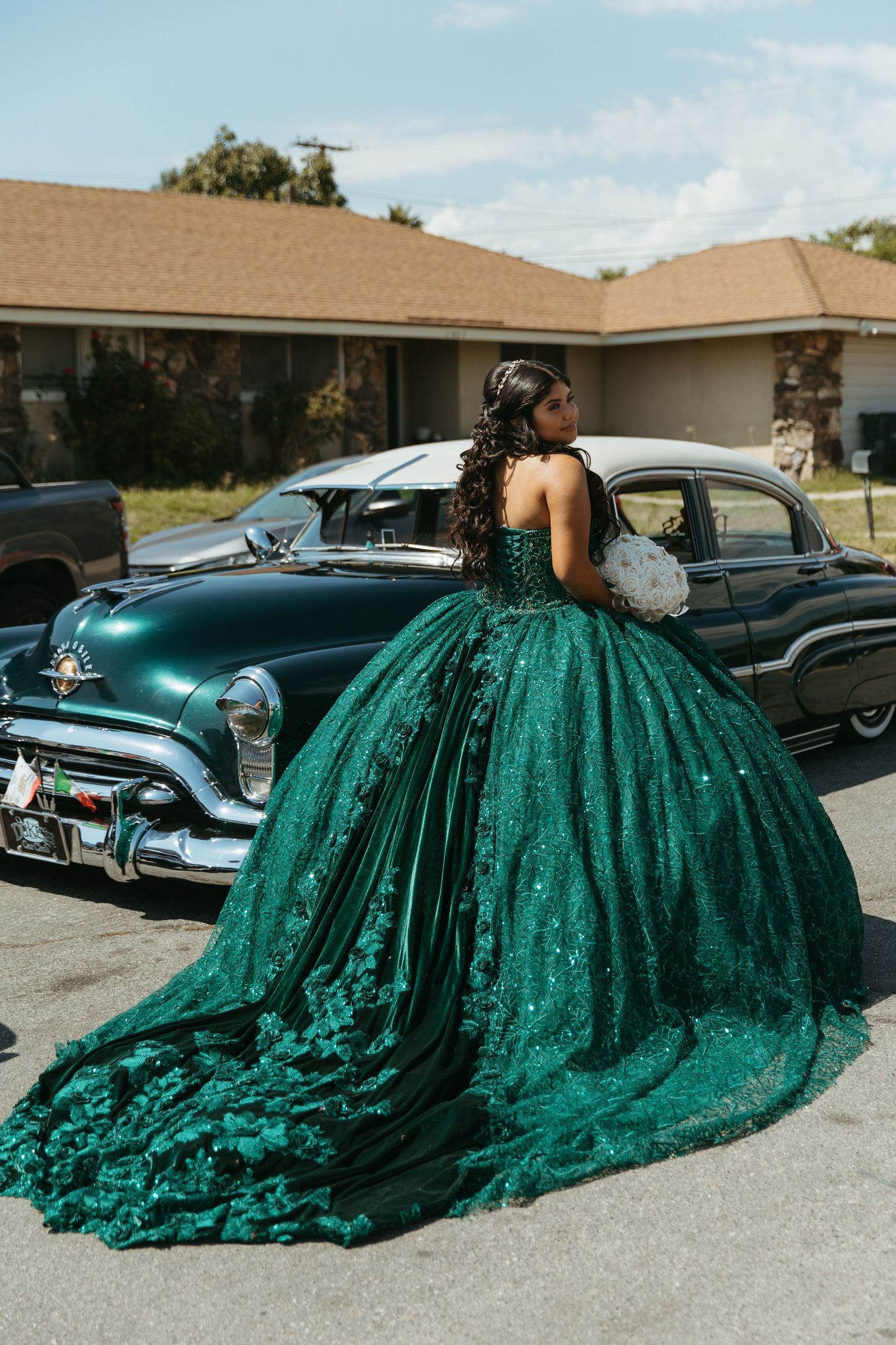 Young woman in emerald green quinceañera ball gown with lace details poses near vintage classic cars.