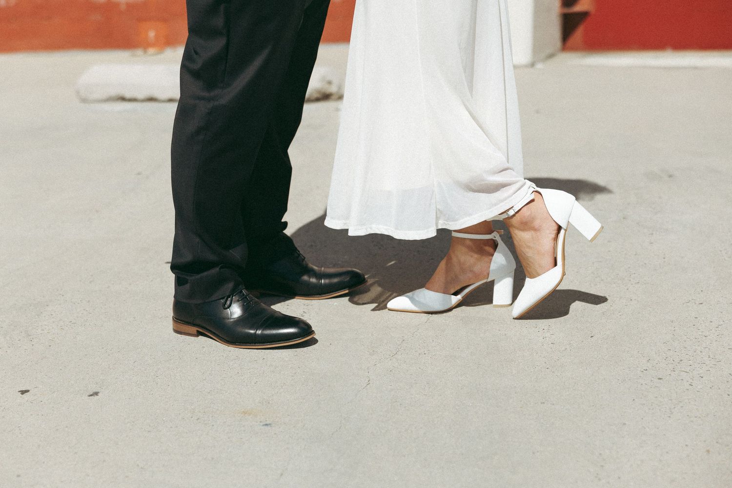 Close-up of bride in white heels and groom in black dress shoes standing together on pavement.