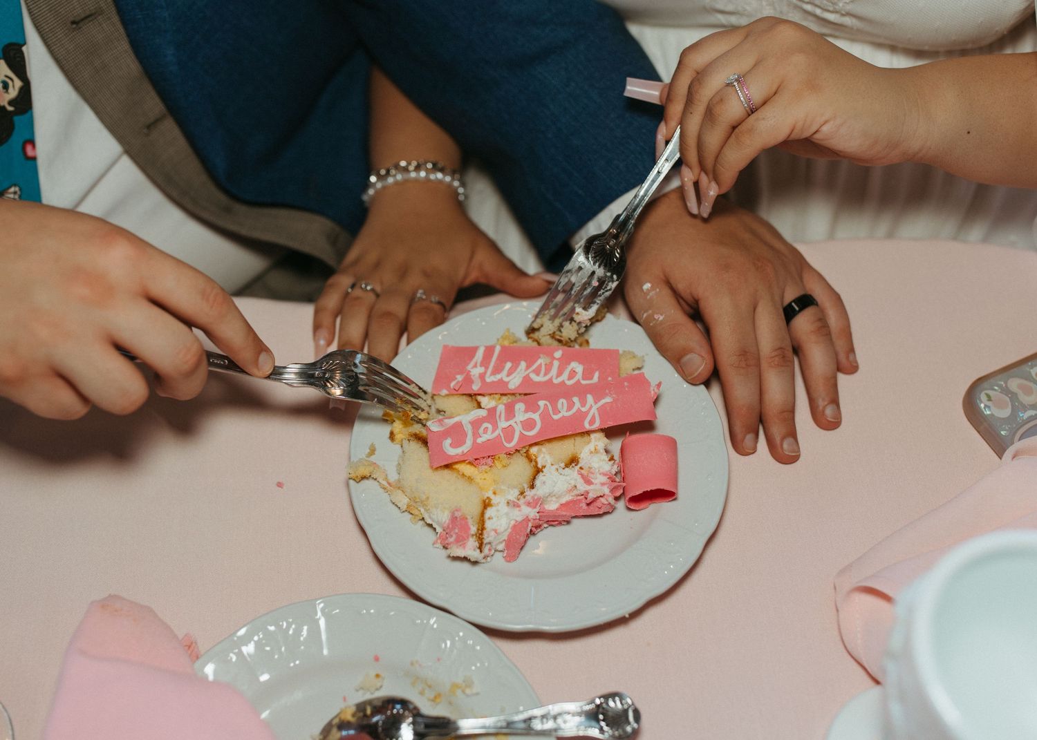 Couple cutting wedding cake on a plate with pink Alycia and Jeffrey name card at reception table.