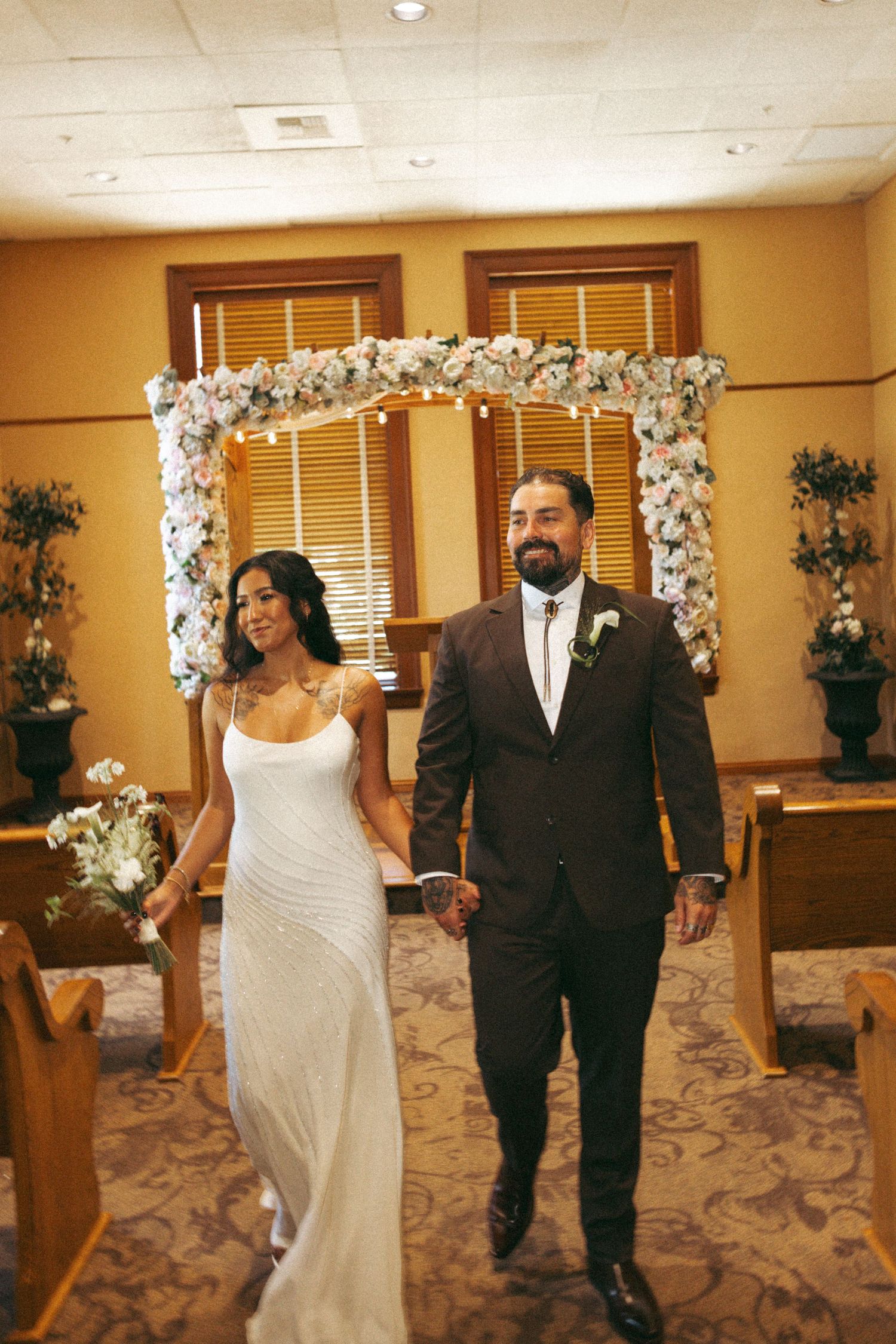 Newlywed couple walks hand-in-hand down the aisle after wedding ceremony beneath a floral arch.