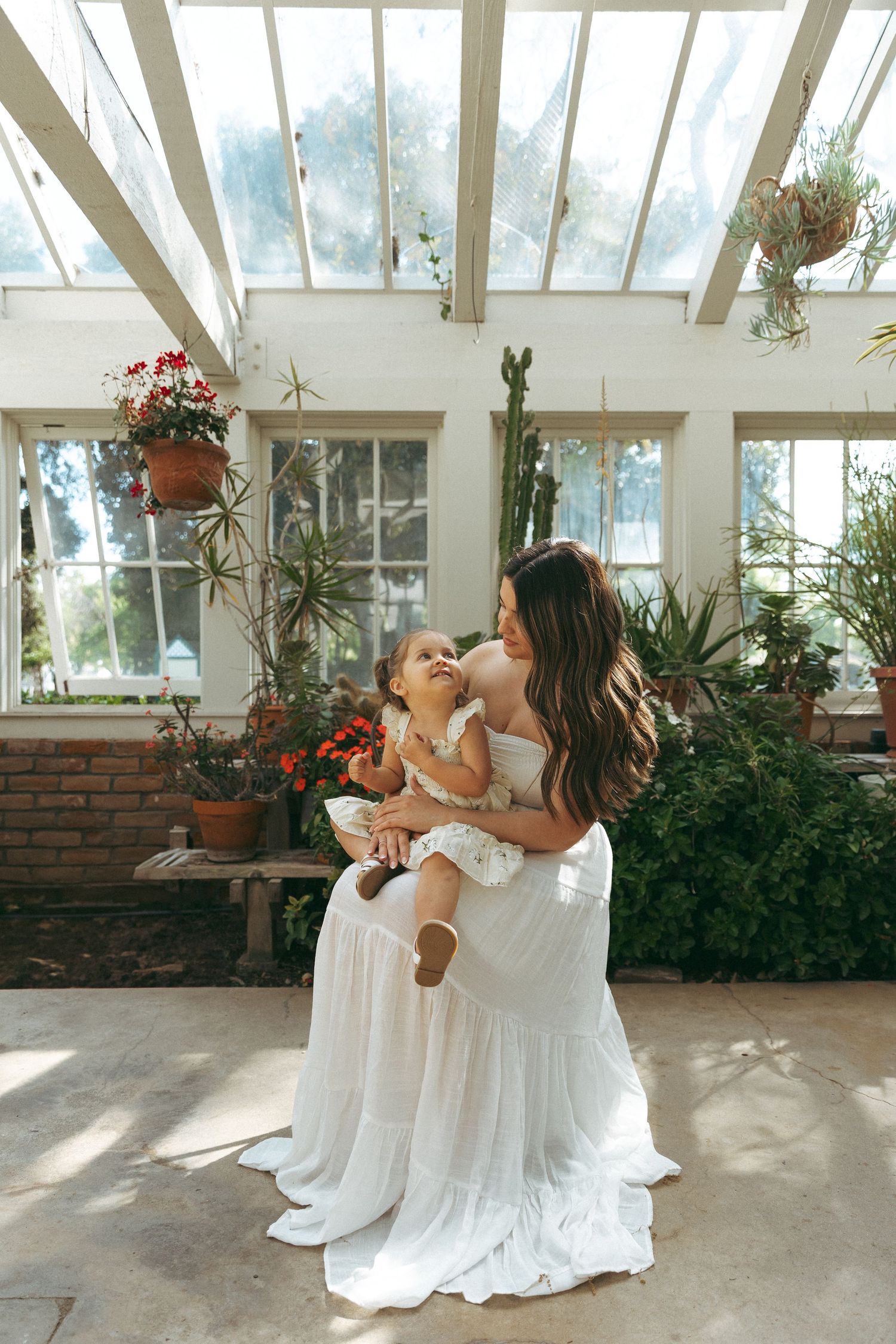 Bride in flowing white gown holds and kisses a young child in a sunlit greenhouse filled with plants.