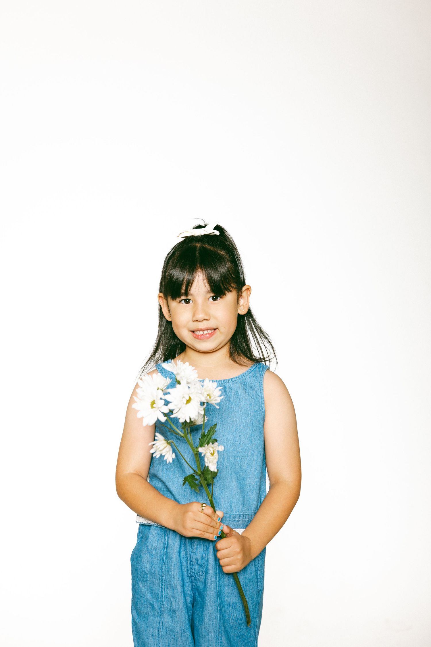Young girl in blue denim jumpsuit holding white daisy flowers, smiling at camera against white background.