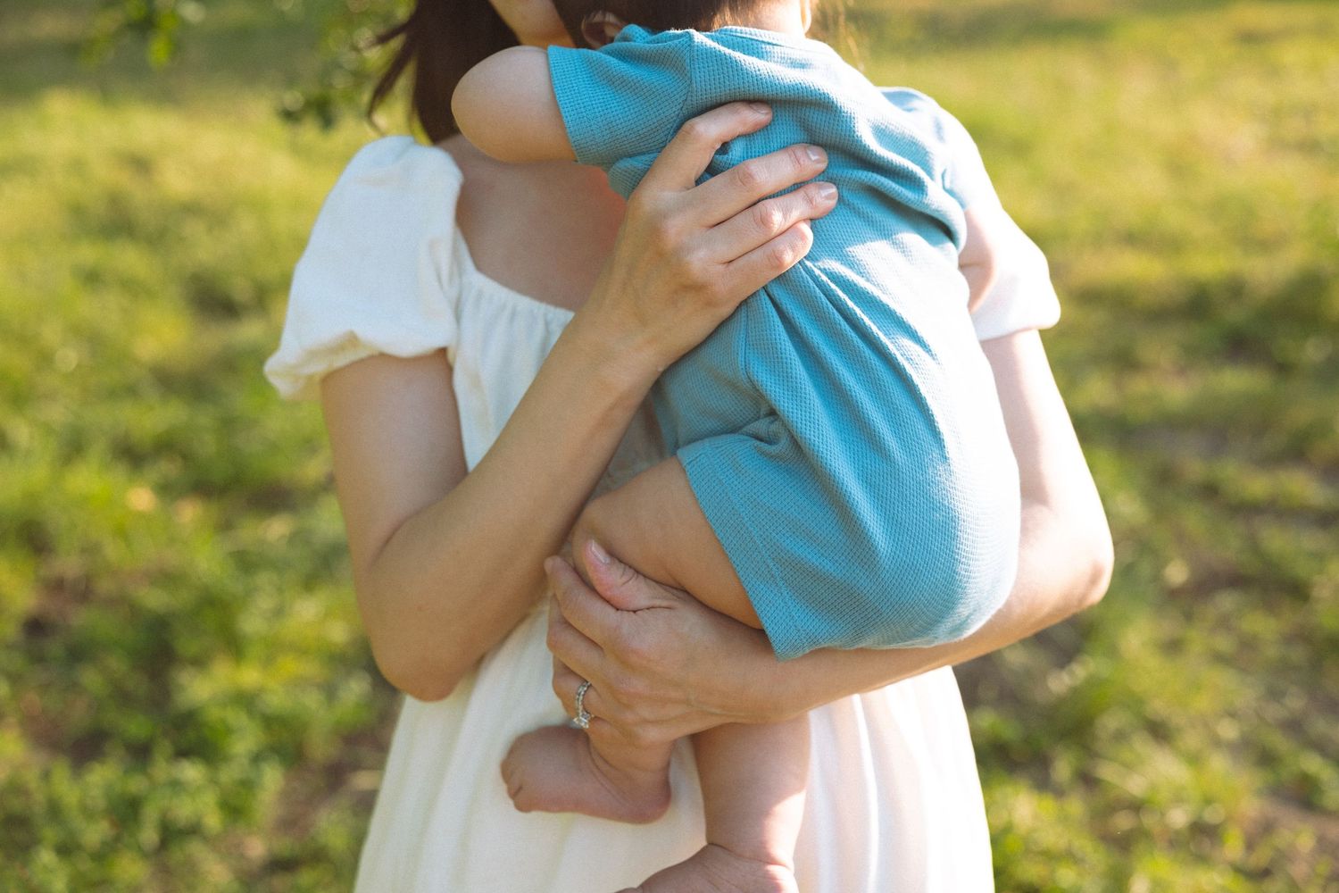 Mother in white dress tenderly holding baby in blue outdoors in sunny green park setting.