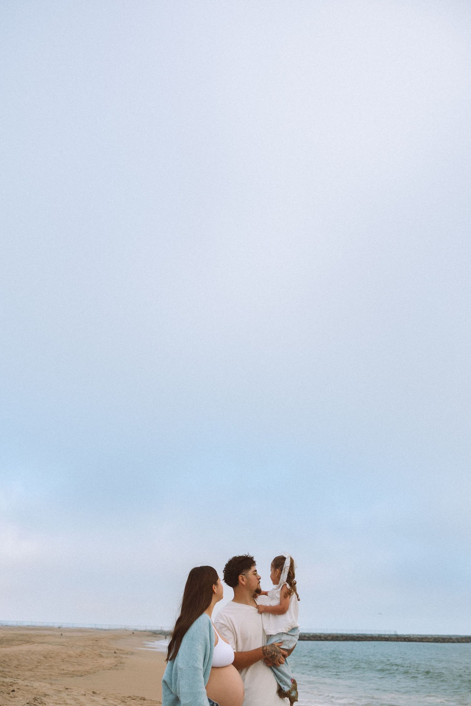Family of three enjoying a peaceful beach day, parents holding their young child with calm ocean waves in the background. Beach Maternity.