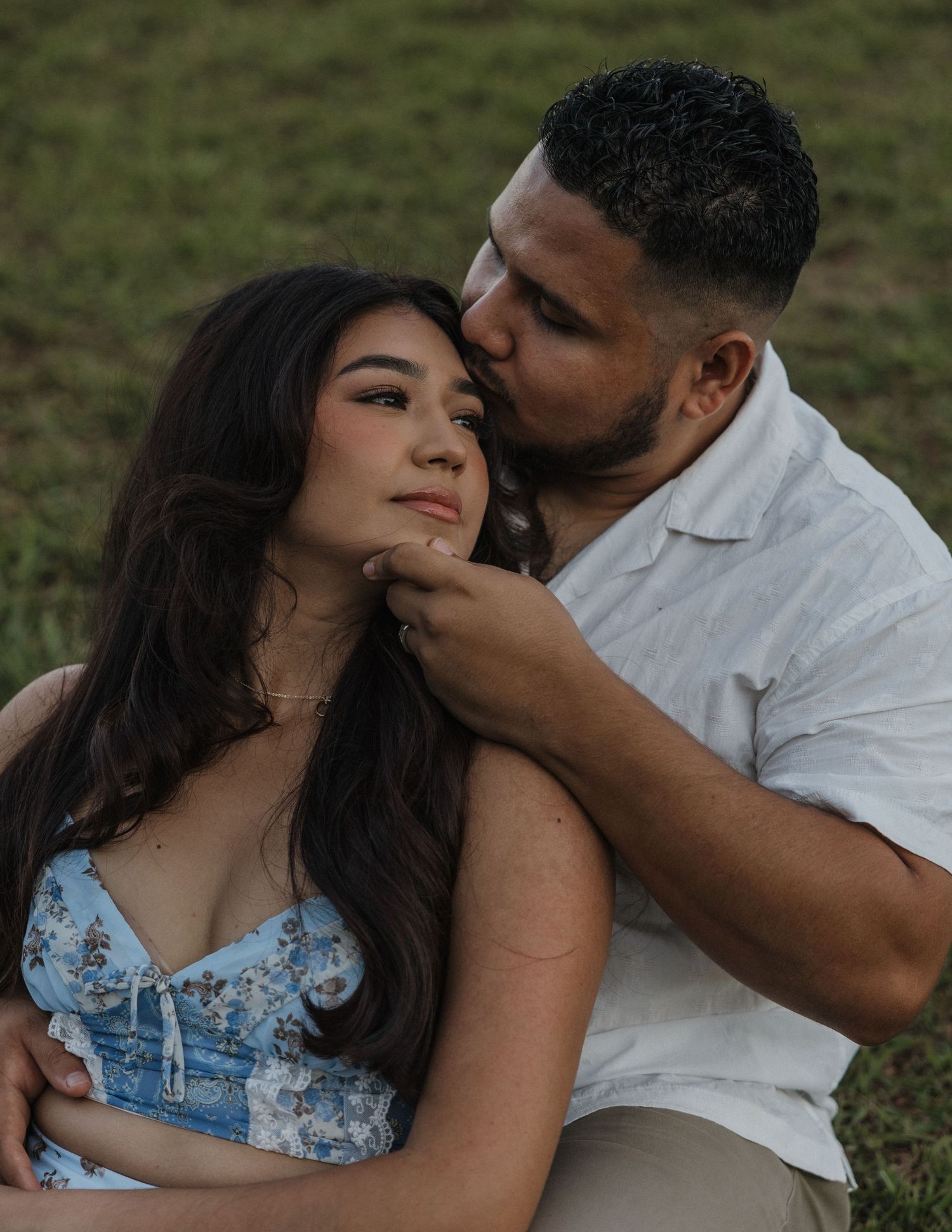 A couple shares a tender moment outdoors, man kissing woman's temple while gently holding her chin.