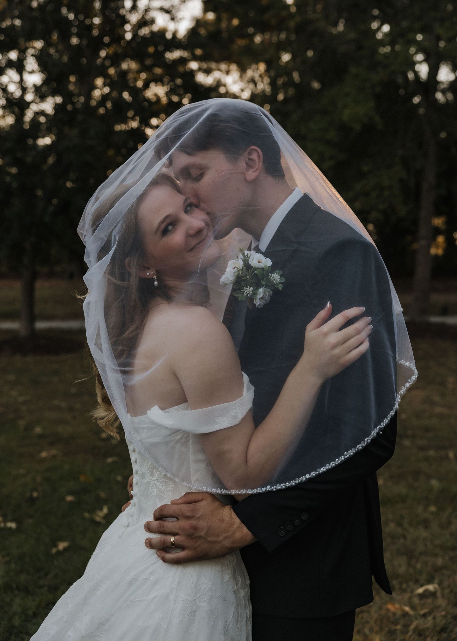 Bride and groom share a romantic kiss under a wedding veil outdoors, surrounded by lush green trees.