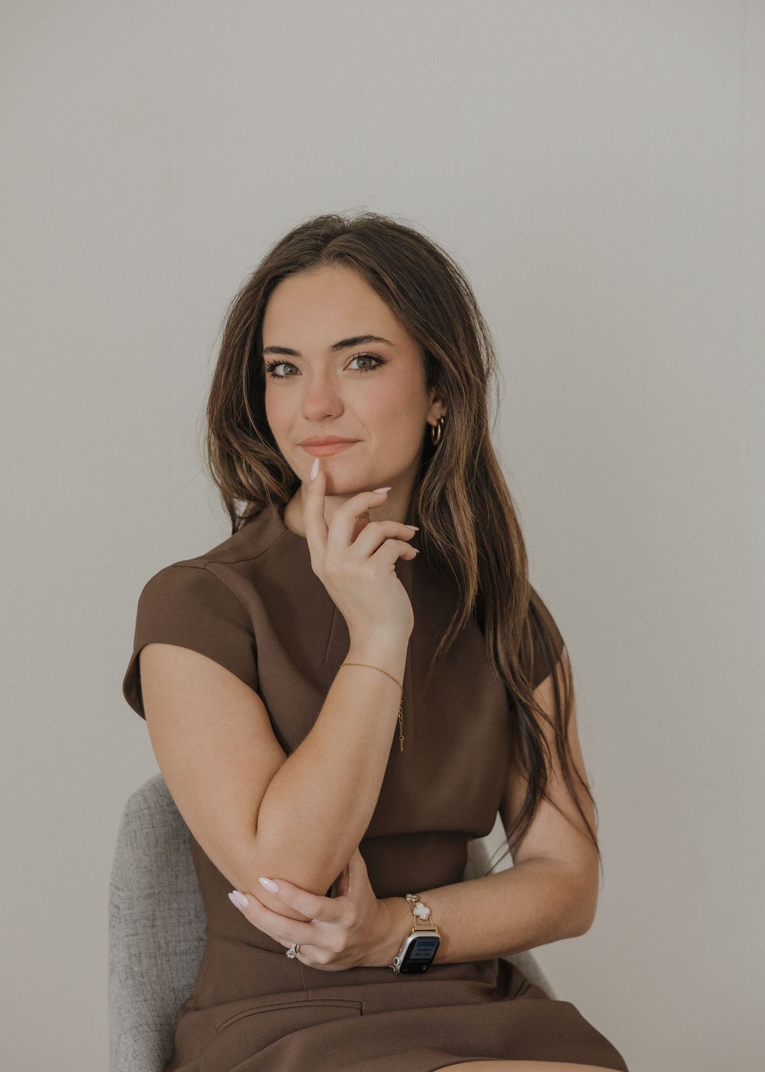 A professional woman in a brown dress sits thoughtfully with her hand on her chin, wearing a gold watch.