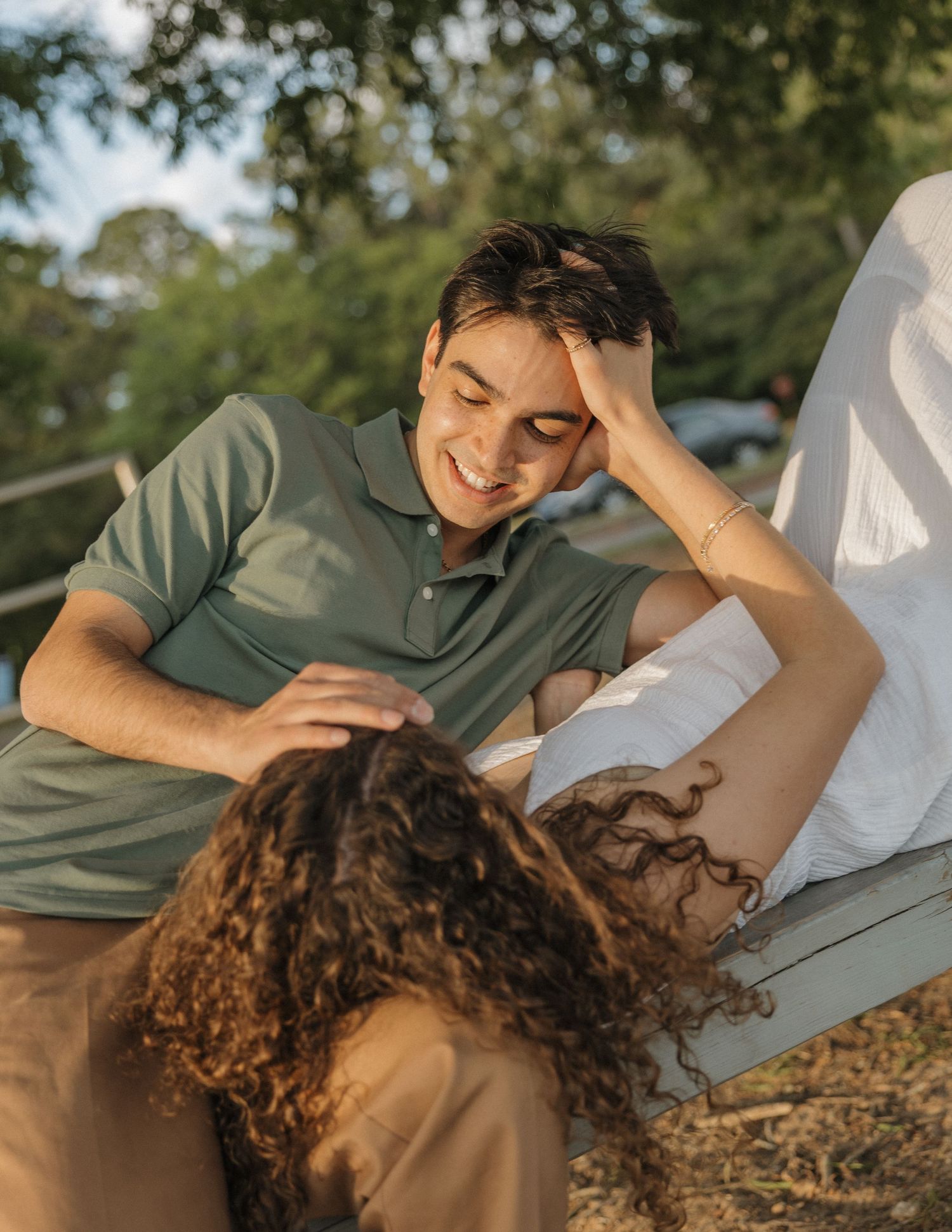 A smiling young man in a green polo shirt relaxes outdoors with a curly-haired woman lying in his lap.