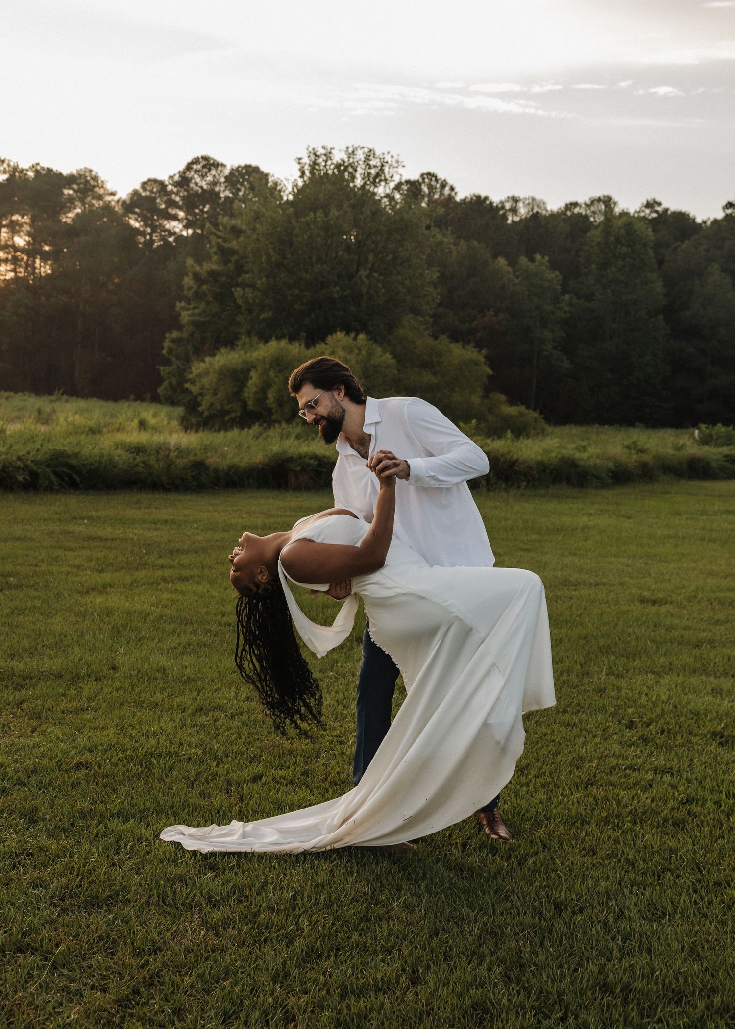 Couple dancing at sunset in a green field, man dipping woman in white dress, trees in background.