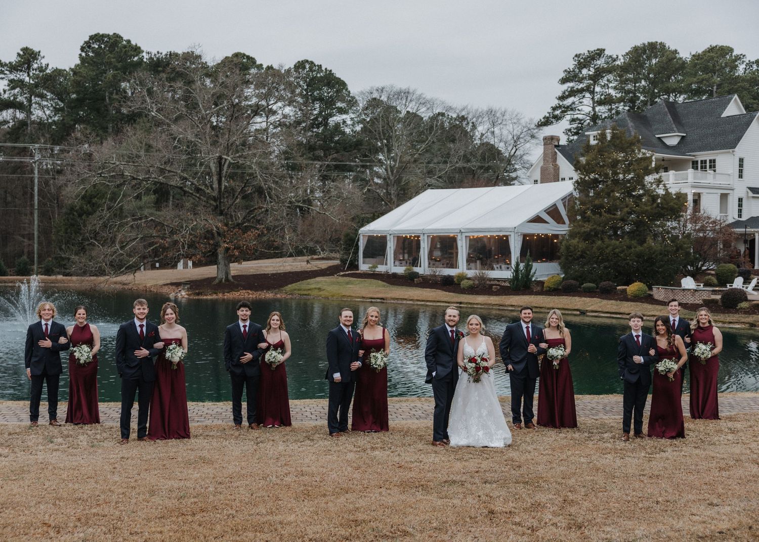 Wedding party posing outdoors near a pond with a white tent venue and trees in the background.