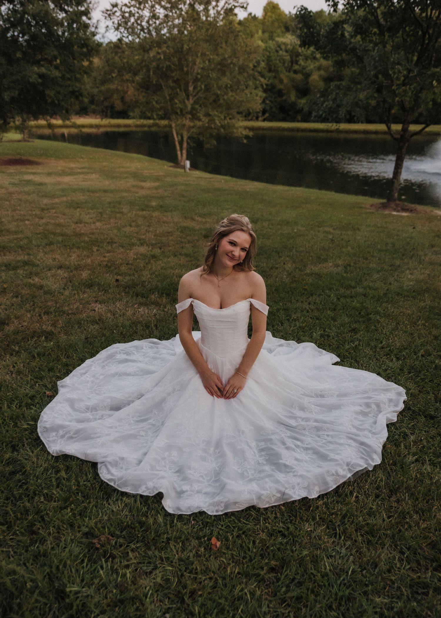 Bride in off-shoulder ball gown wedding dress sitting on green grass with pond and trees in background.