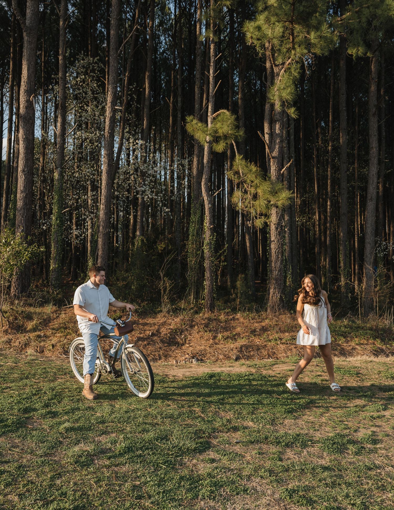A couple enjoys a sunny day in a pine forest, with a man walking a bicycle and a woman in a white dress smiling nearby.