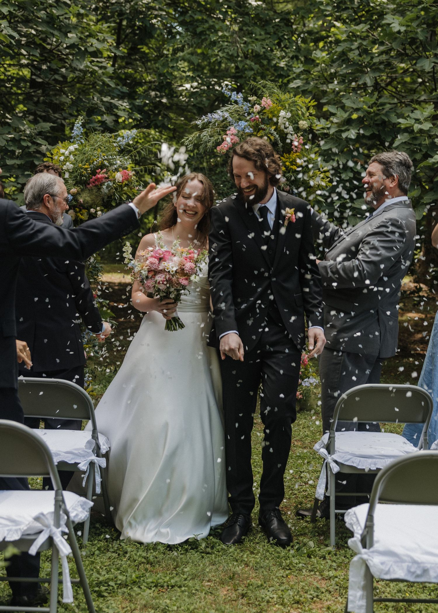 Bride and groom walk down aisle as guests toss white confetti at outdoor garden wedding ceremony.