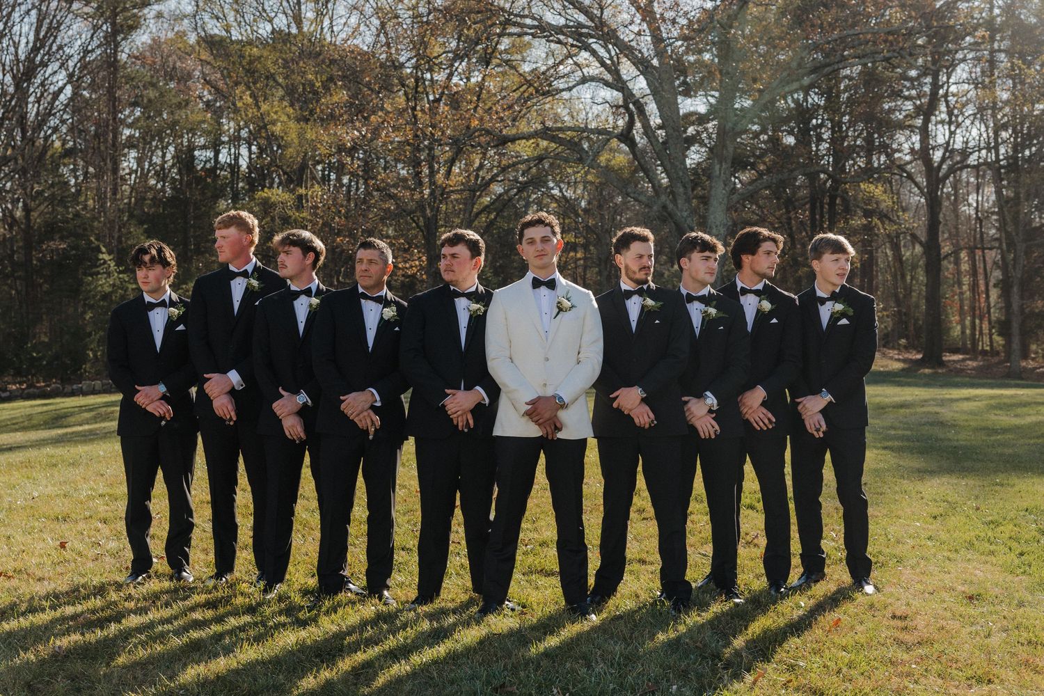 Groomsmen in black tuxedos and one in white stand together for formal wedding photos on a sunny autumn day.