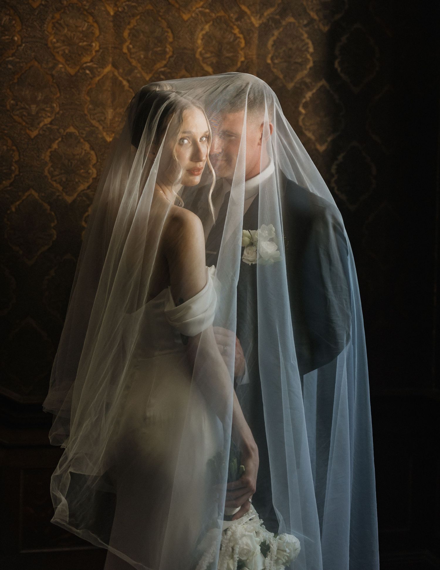 Bride and groom share an intimate moment together under a flowing white wedding veil against a golden damask backdrop.